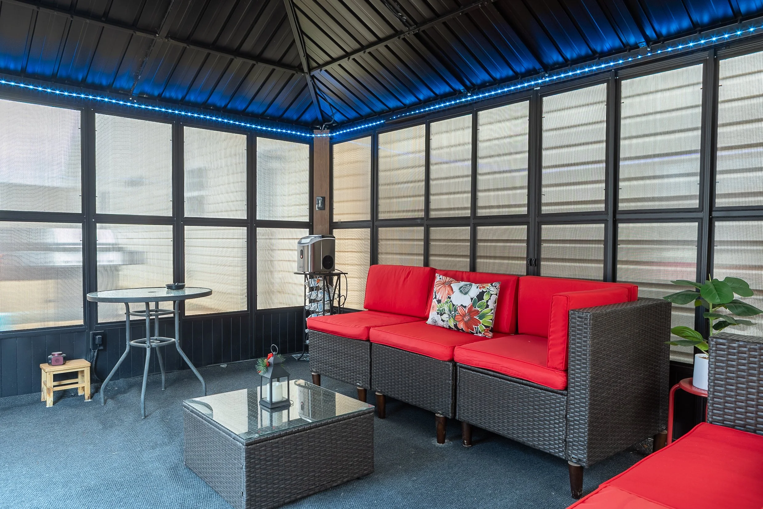 Indoor patio area with black wicker furniture, red cushions, a glass-top coffee table, small side tables, and potted plants, illuminated by blue string lights along the ceiling.