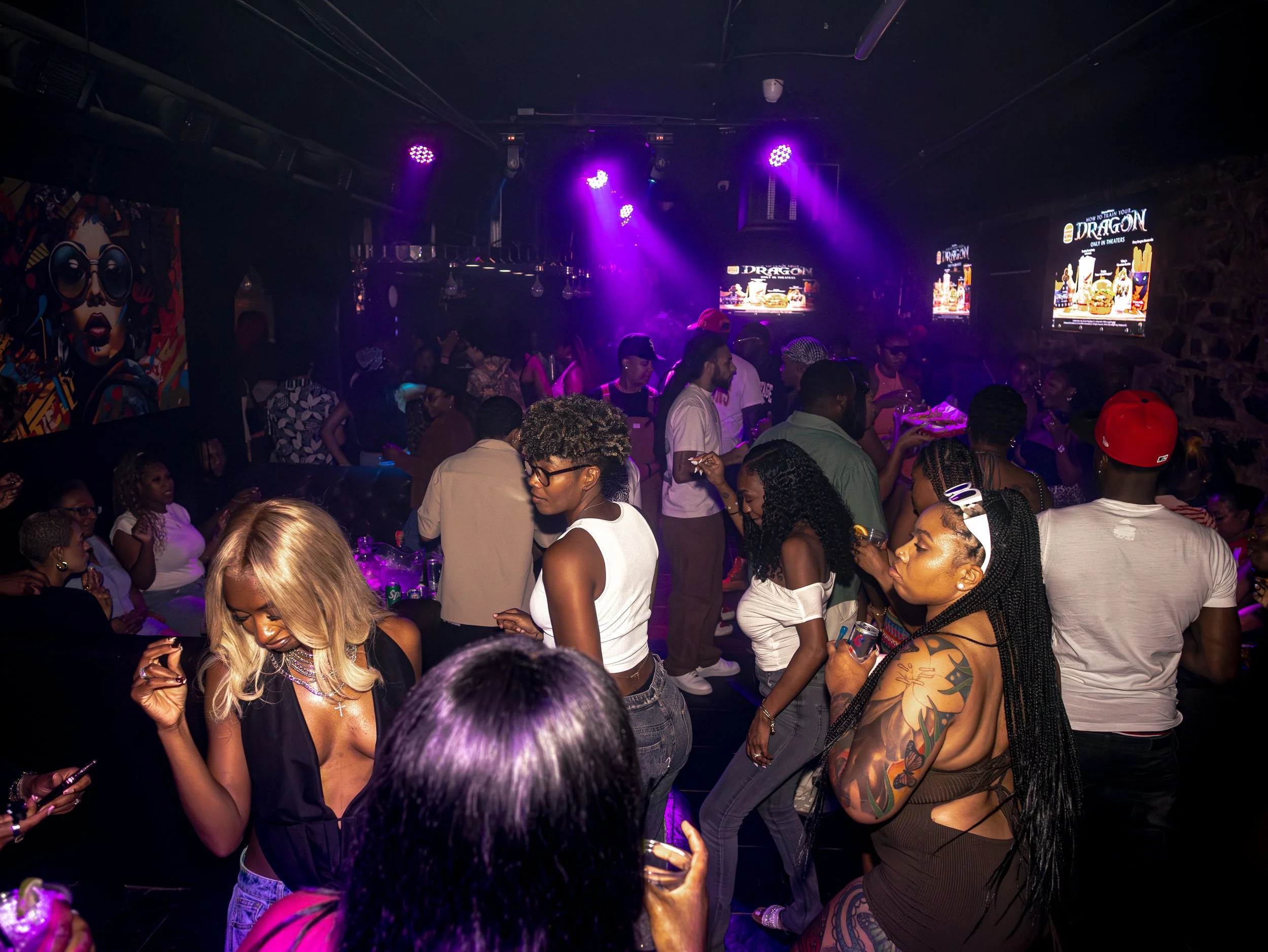 People dancing and socializing in a nightclub with purple lighting, some looking at their phones, and two TVs displaying promotional content in the background.