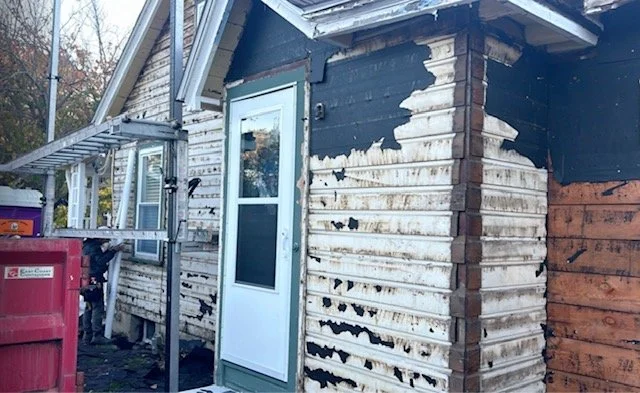 Side of a house with peeling paint and visible damage to the siding and wall. There is a door with a window and some outdoor equipment including a ladder and a purple trash bin.