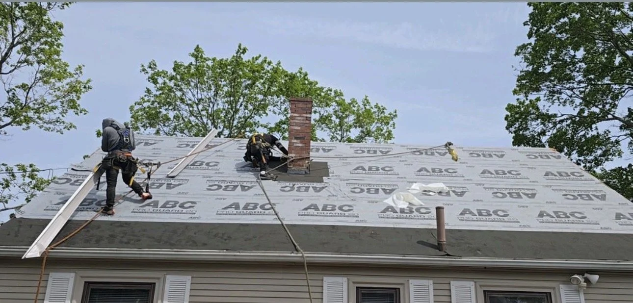 Two workers on a house roof installing a weather-resistant barrier. The roof has a brick chimney and various ventilation pipes. The workers are wearing safety harnesses and tools.