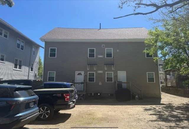 A three-story residential building with gray siding and multiple windows, situated on a dirt lot with parked cars in the foreground.