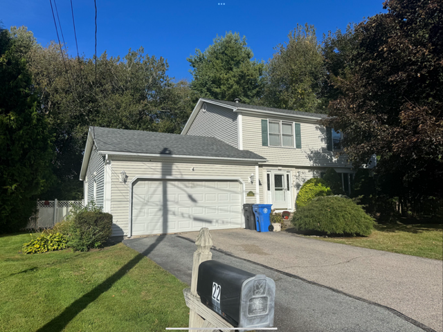 A two-story house with gray siding, a double garage, and a driveway, surrounded by trees and greenery on a sunny day.