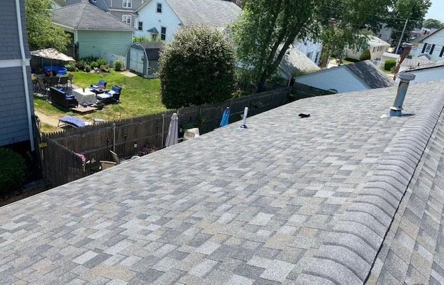 View of gray shingled roof with vents, overlooking a backyard with trees, grass, and outdoor furniture.