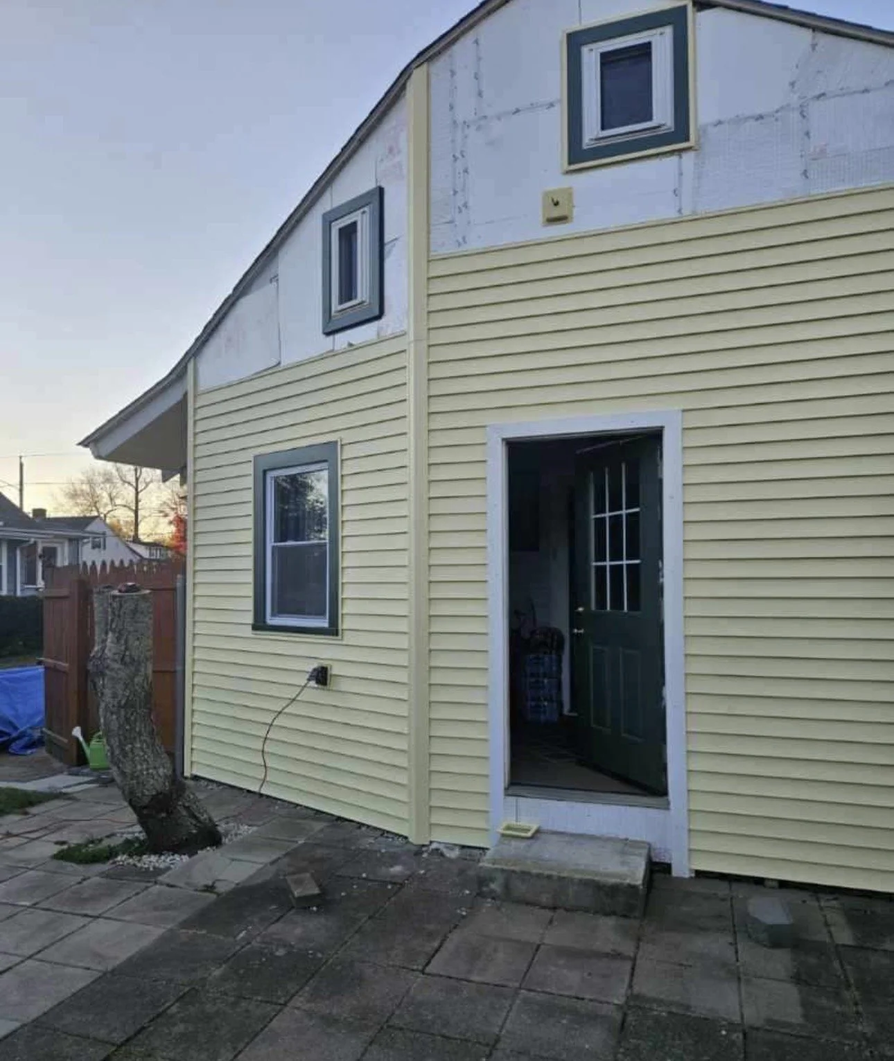 A house under renovation with yellow siding and two windows on the upper level. The lower level has a window and a door with a small step leading outside. A tree stump is visible near the house, and the backyard has a paved patio area.