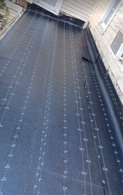 Black roofing membrane with construction markings and measurements, on a building rooftop before installing decking or shingles.