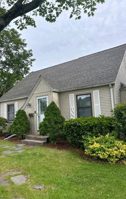 A house with beige siding, a gray roof, and white shutters, surrounded by bushes and a lawn with a stone pathway.