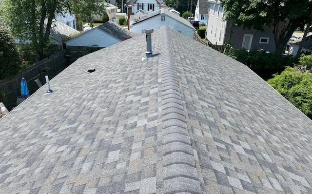 Aerial view of a house roof covered in gray asphalt shingles, with vents and a chimney pipe, surrounded by neighboring houses and trees.