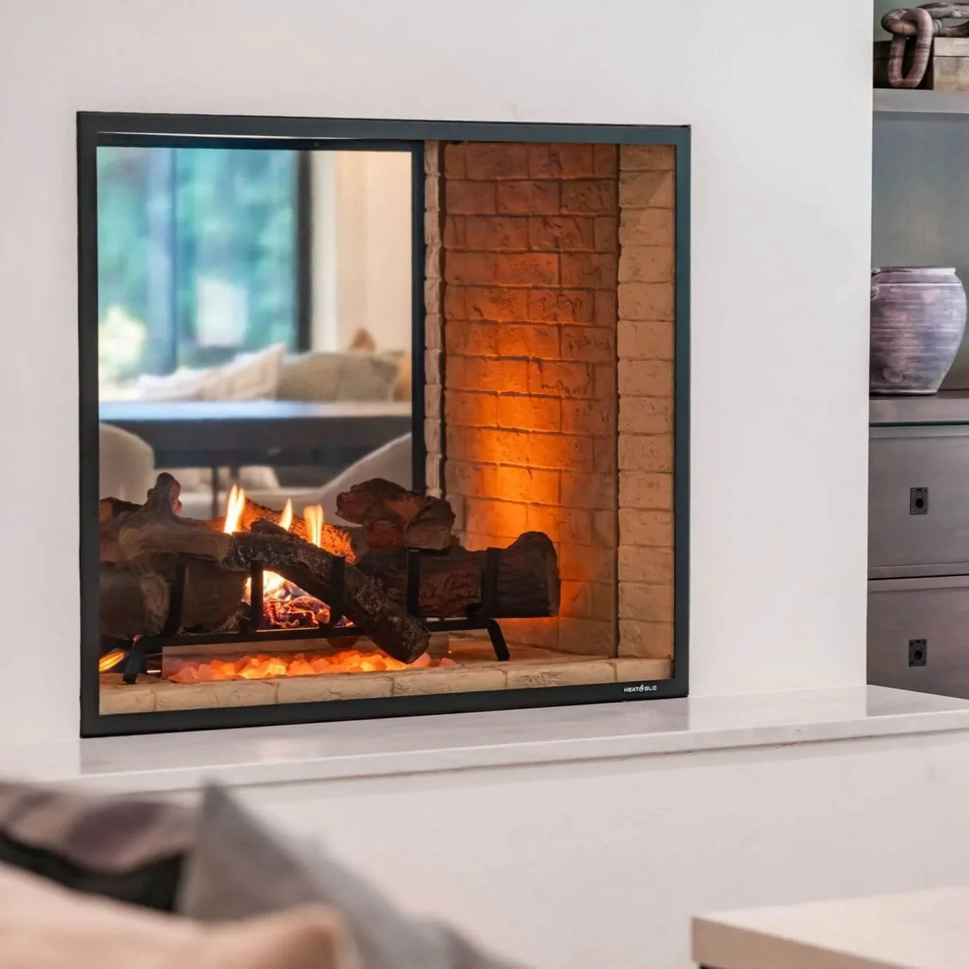 A modern indoor fireplace with realistic-looking logs and flames, and a mirror reflecting a cozy living room with beige sofas and large windows.