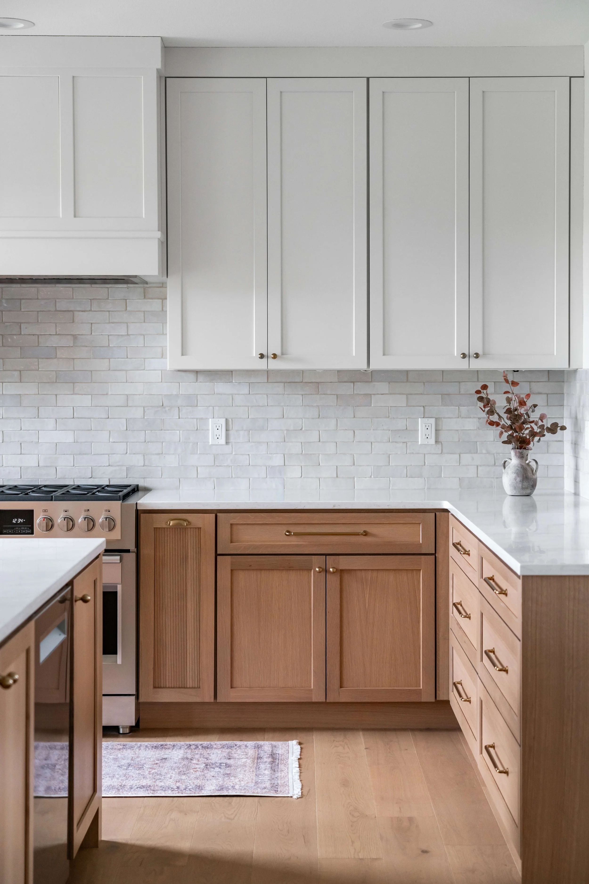 A modern kitchen with white upper cabinets, light brick backsplash, and natural wood lower cabinets. There is a countertop with a vase of dried leaves on it.