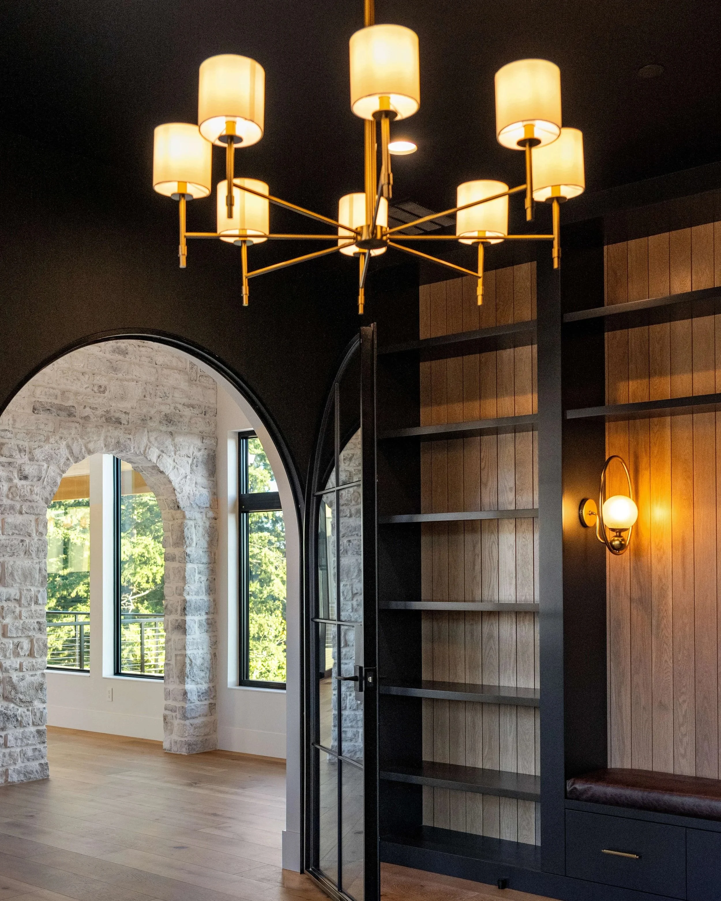 Interior of a modern room featuring a black and gold chandelier, a built-in black and wood bookshelf, a wall-mounted lamp, and arched stone doorway leading to a bright room with large windows and trees outside.