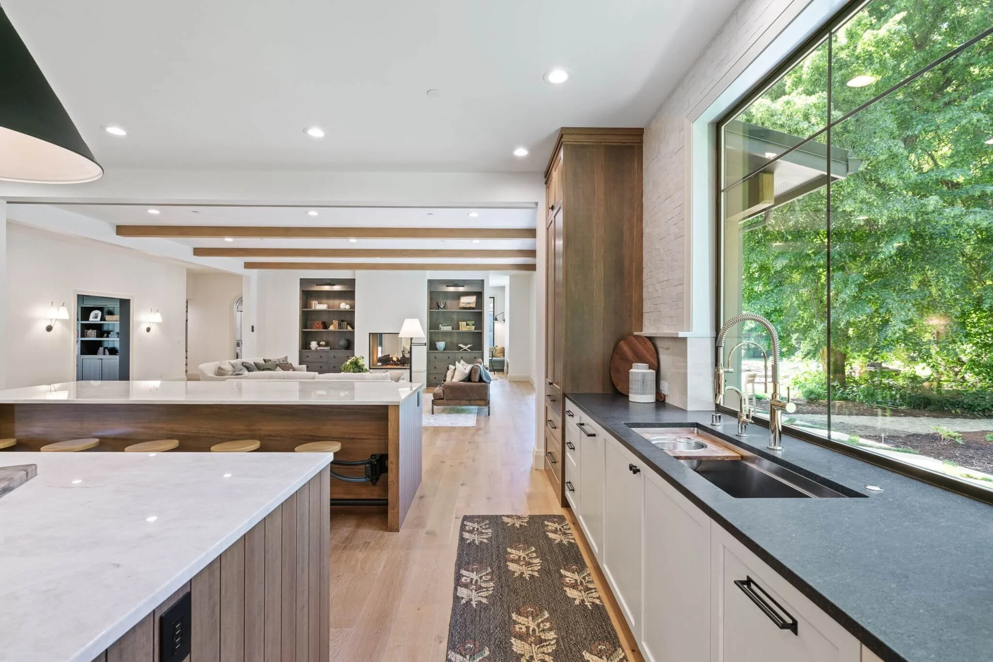 Open-concept kitchen with white cabinets, black countertop, large window, and view of green trees outside, part of a spacious living area with wooden beams and bookshelf.