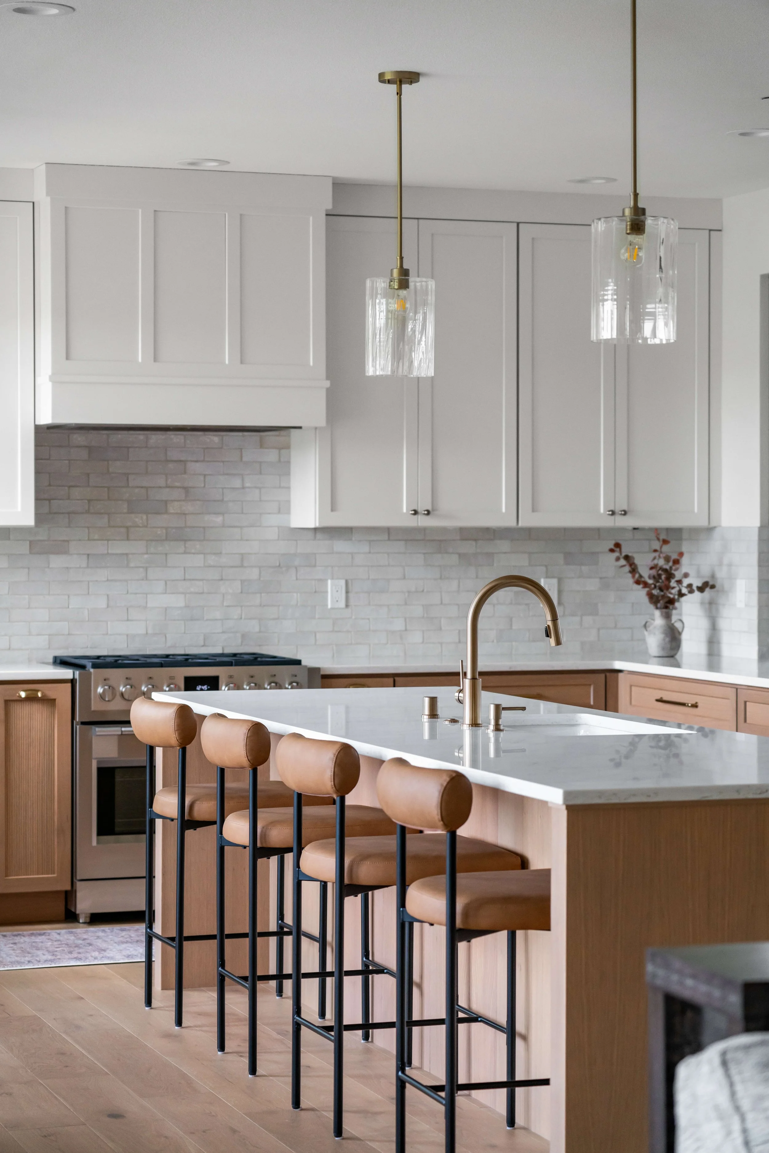 Modern kitchen with white cabinets, a gray brick backsplash, wooden lower cabinets, a marble countertop island with a gold faucet, and four tan barstools.