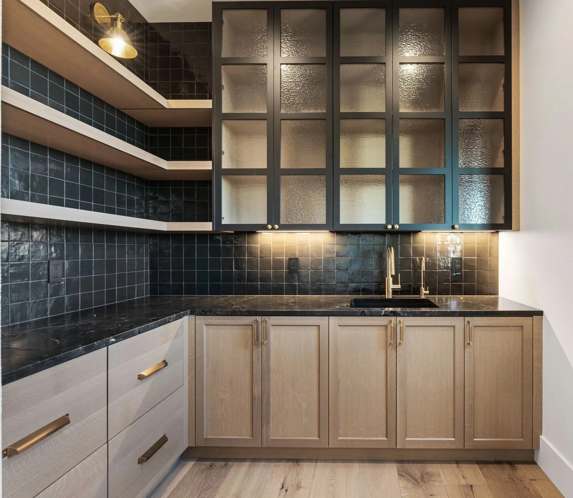 A modern kitchen with black tiled backsplash, beige cabinetry with gold hardware, black countertops, glass-front upper cabinets, and a single sink with a gold faucet.