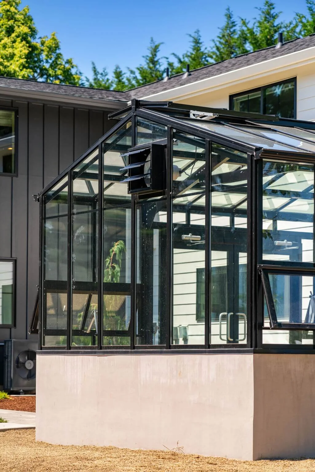 A modern glass greenhouse attached to a house with dark and white siding, set outdoors with trees and a clear blue sky in the background.