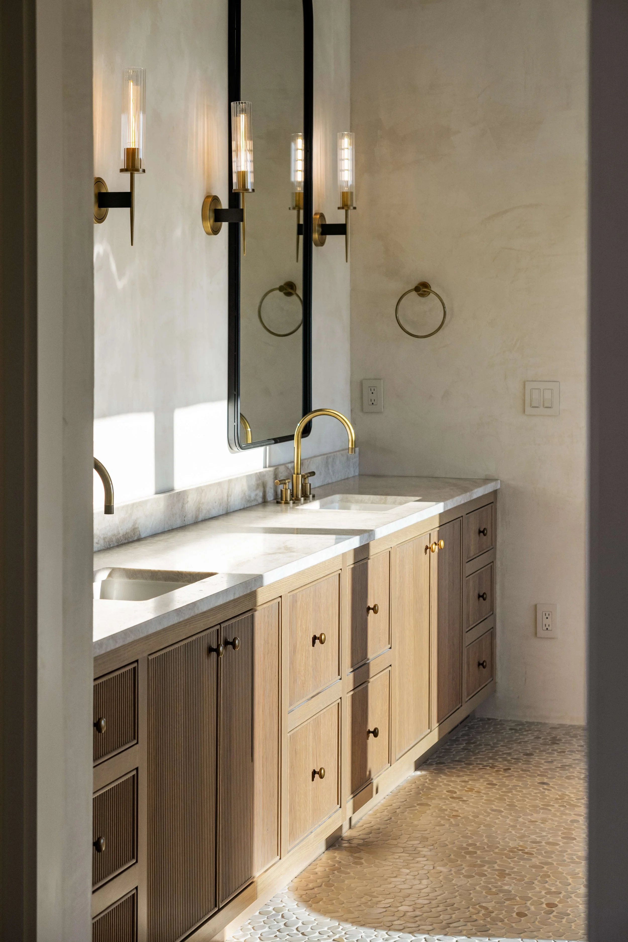 A modern bathroom vanity with wooden cabinets, a white marble countertop, two sinks, and gold fixtures. A large mirror and wall-mounted gold light fixtures are above the sinks. The wall is painted a light neutral color.