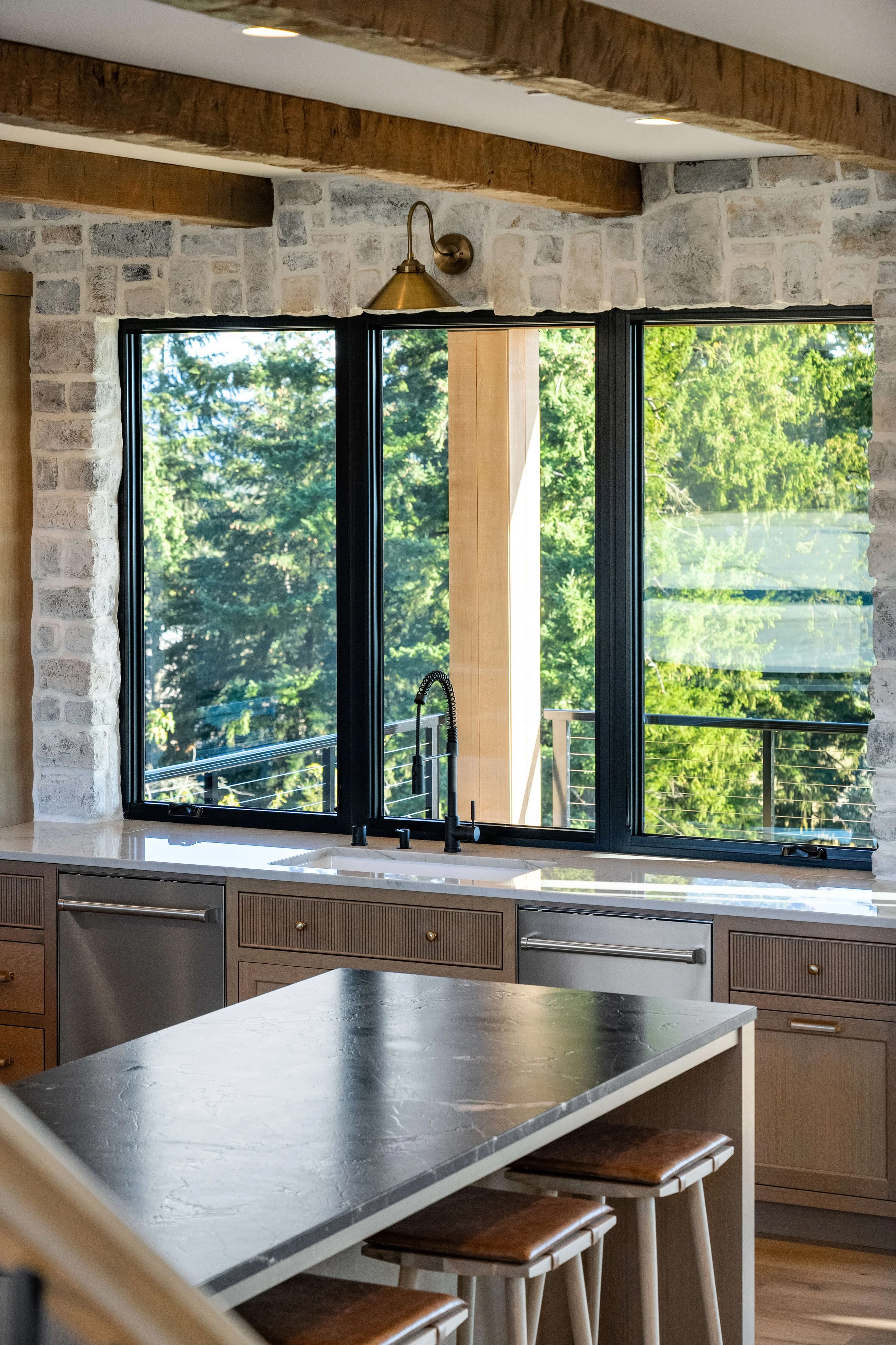 Modern kitchen with large window overlooking green trees, featuring a black sink with a black faucet, light wood cabinets, and a black countertop island with wooden stools.
