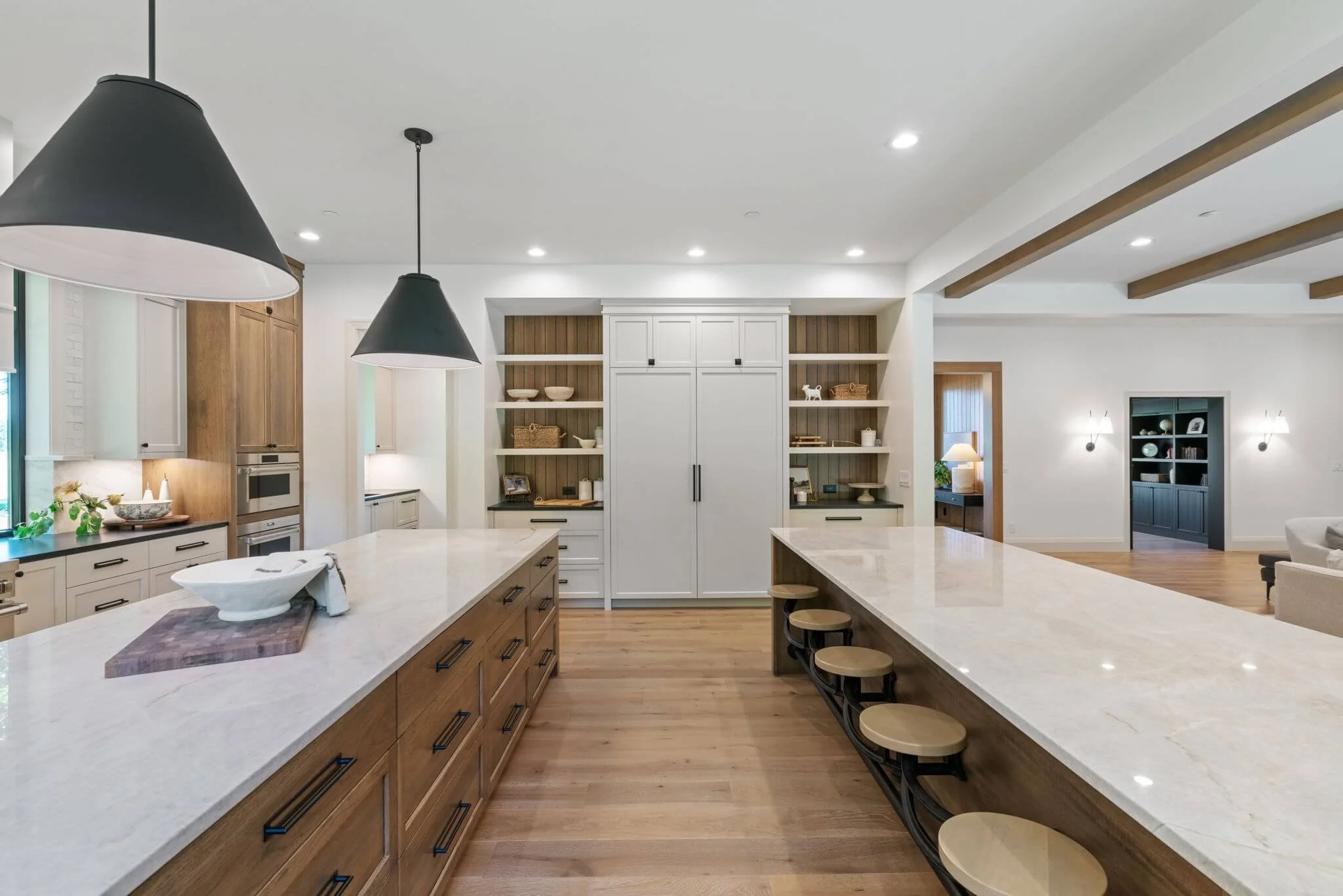 Modern kitchen with white and wooden cabinets, large marble island with bar stools, pendant lights, hardwood floors, built-in shelves, and adjacent living area.