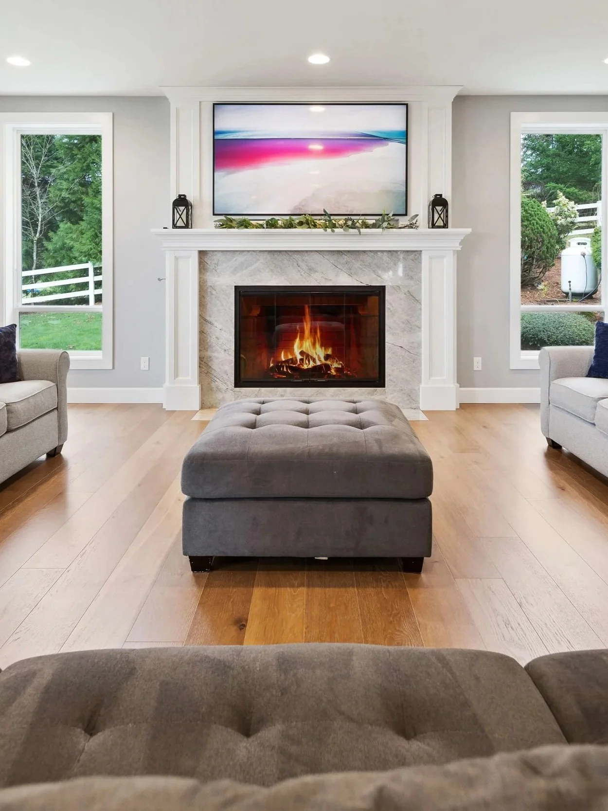 Living room with a fireplace, a large TV above the mantel, two windows on either side showing greenery outside, and an ottoman in the center.