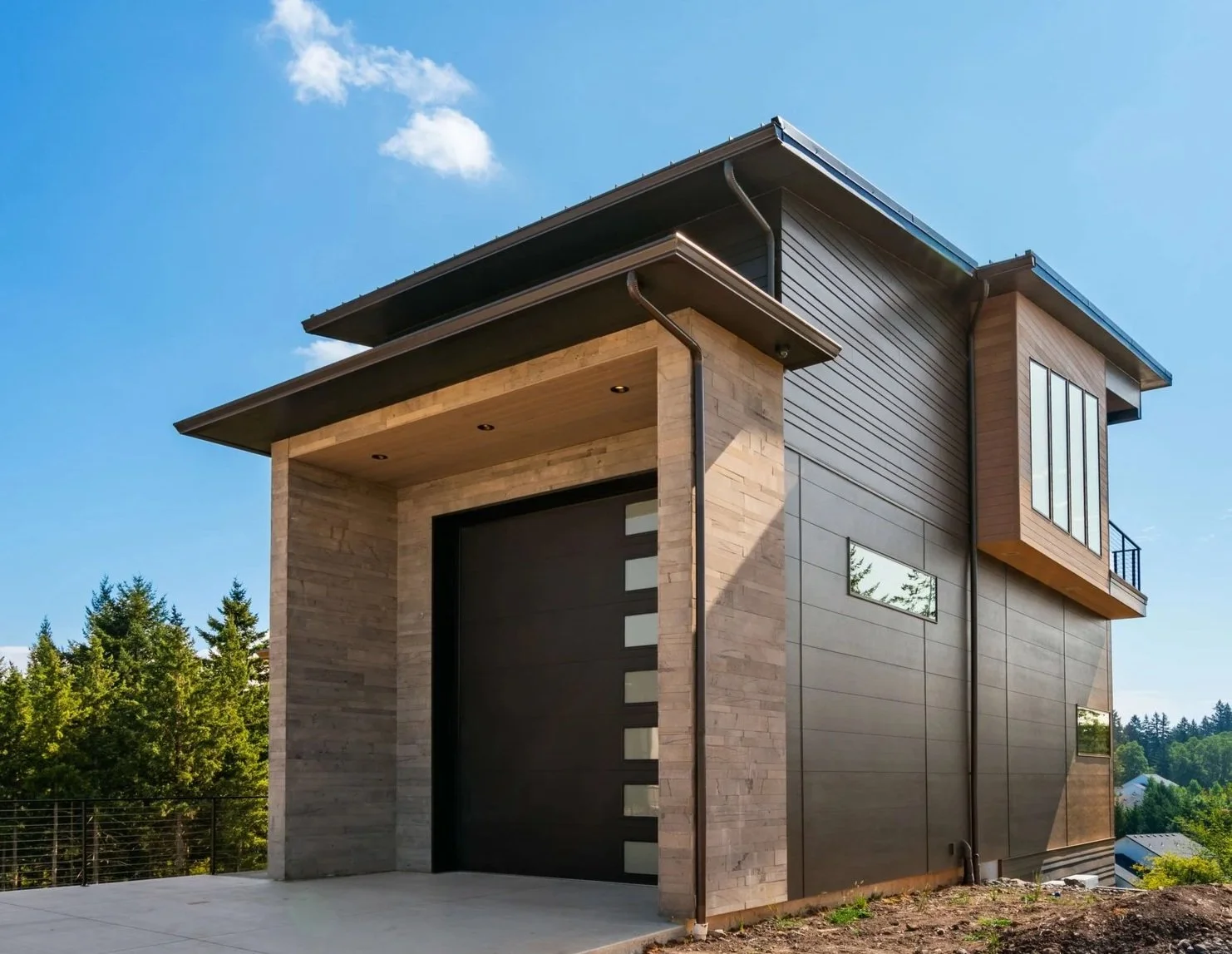 Modern custom shop in Camas, WA with a flat roof, large windows, and a sleek black garage door, set against a backdrop of trees and a blue sky with a few clouds.