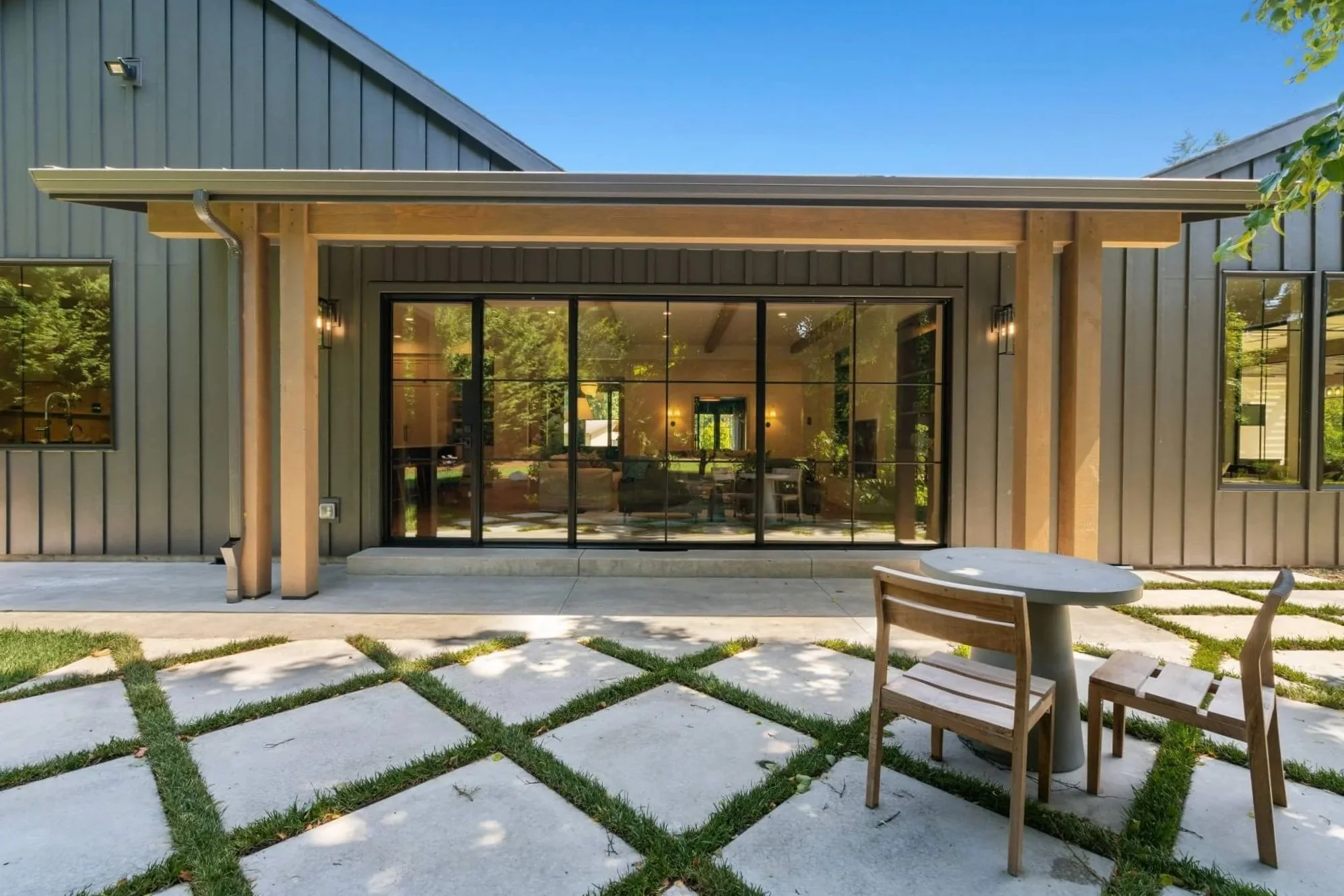 Modern house with large glass sliding door, wooden patio furniture, and a grid-patterned concrete patio with grass.