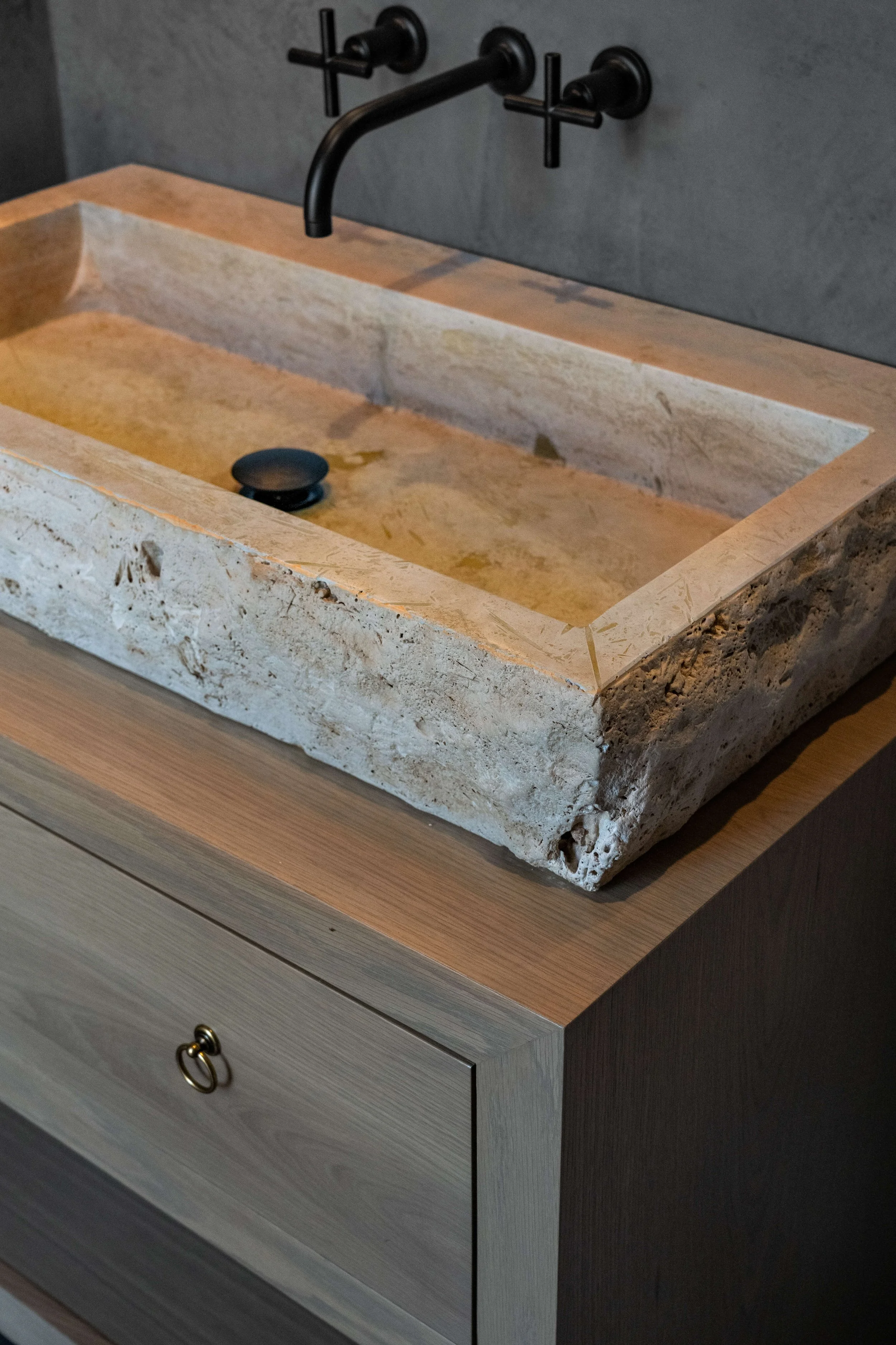 A rustic stone sink with a black drain, mounted on a wooden cabinet, with a black wall-mounted faucet above it.