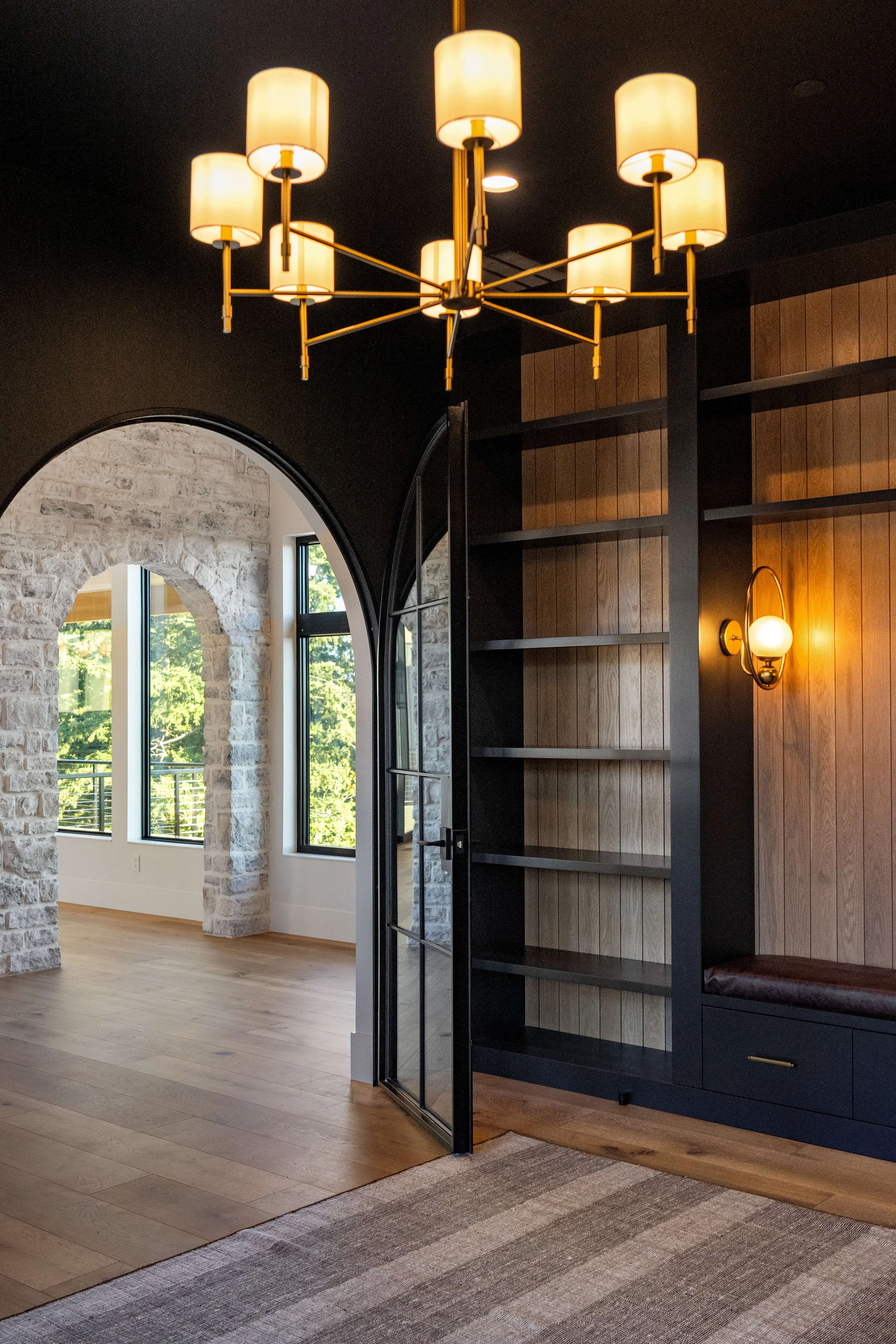Interior of a room featuring a modern chandelier with multiple lamps, a black built-in bookshelf with wooden panels, a stone archway, large windows with a view of trees, and a wall sconce with a globe-shaped light.