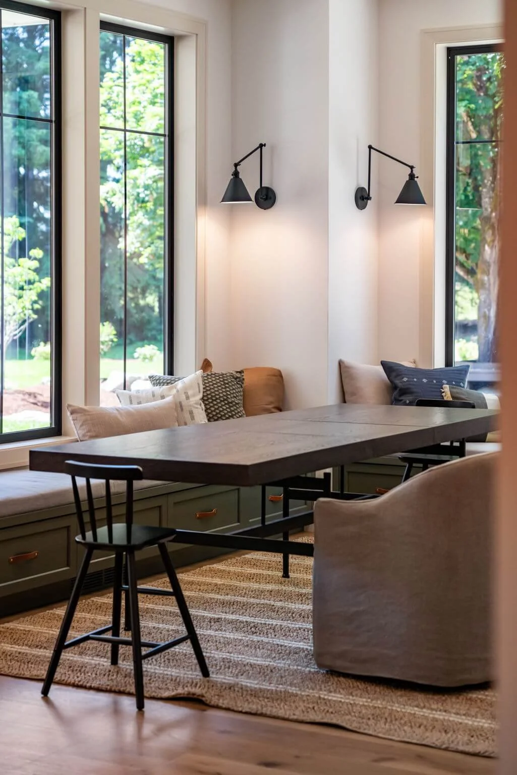 A cozy corner of a room with a wooden table, black chair, a built-in window seat with cushions, and two wall-mounted black lamps, with large windows showing greenery outside.
