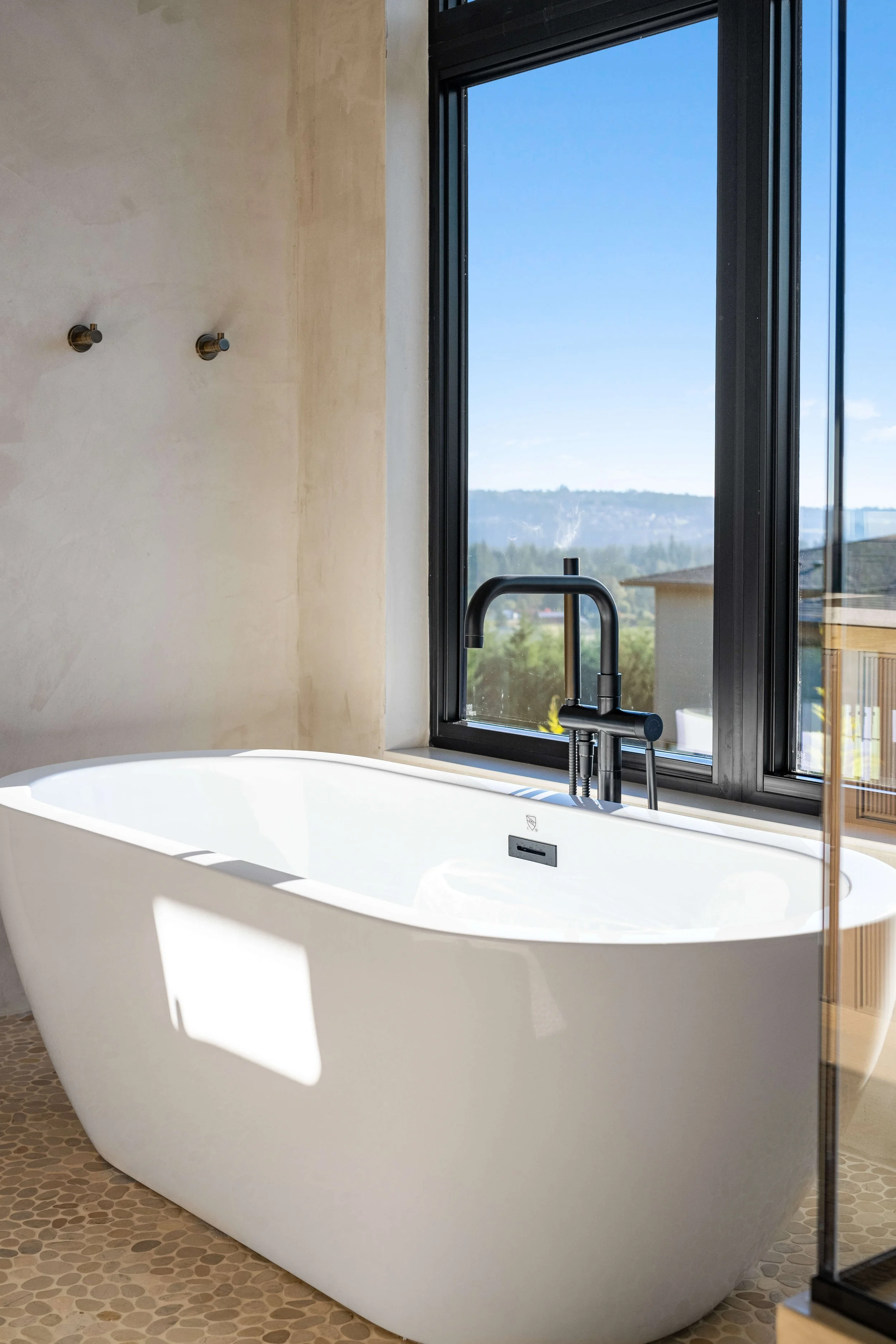 Modern freestanding bathtub next to a large window with scenic outdoor view, black faucet, and a pebble-textured floor.