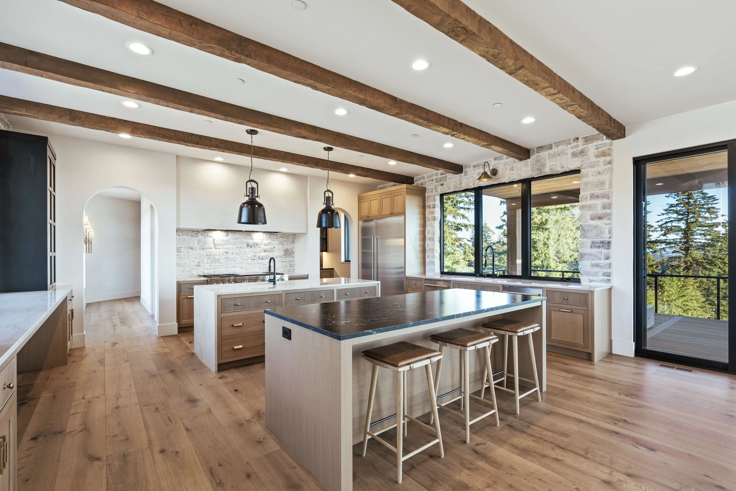 Modern kitchen with wood beams on ceiling, large windows showing trees outside, island with dark countertop, bar stools, and stainless steel appliances.