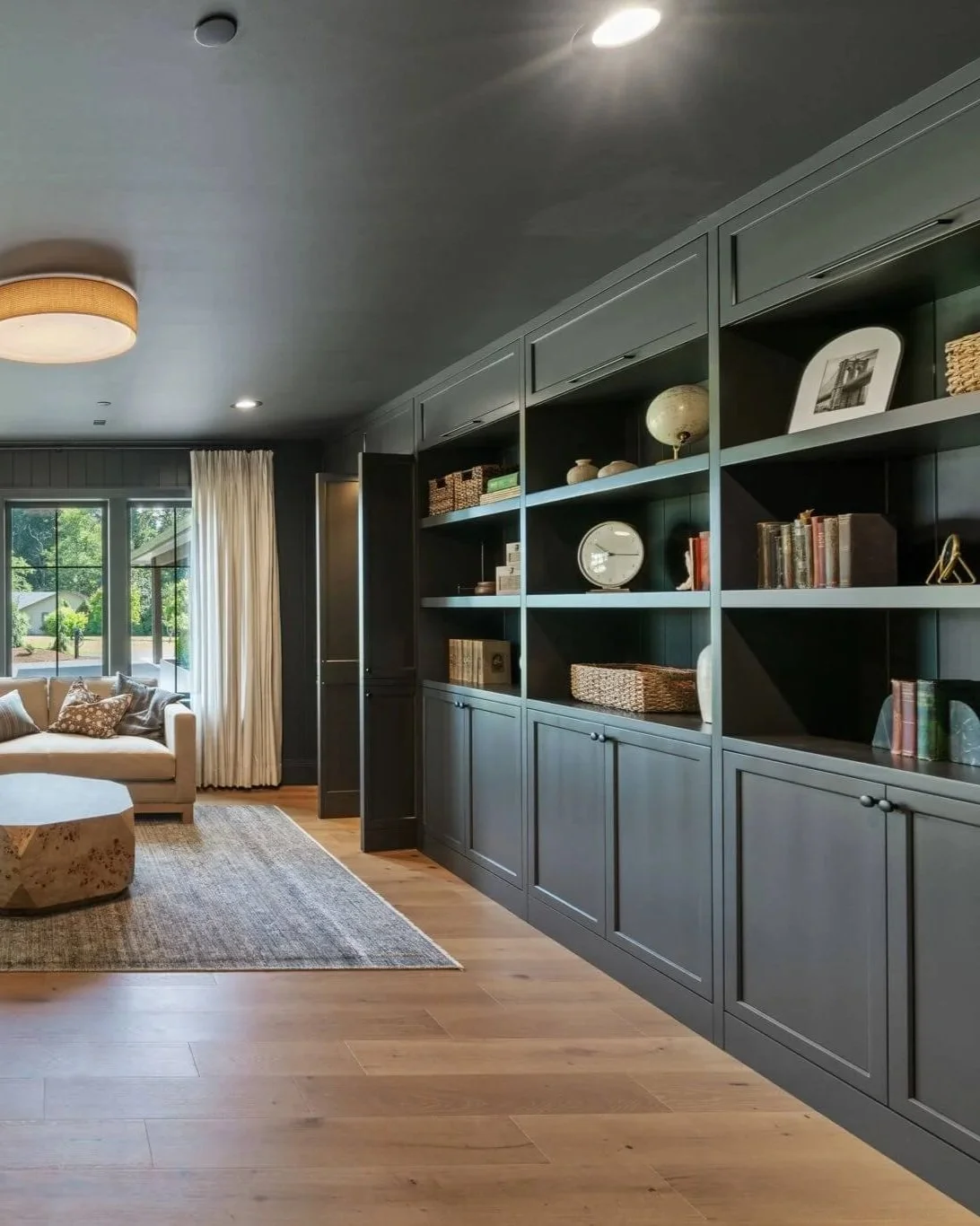 Living room with dark gray built-in shelves, beige sofa near window with curtains, wooden floor, and coffee table.