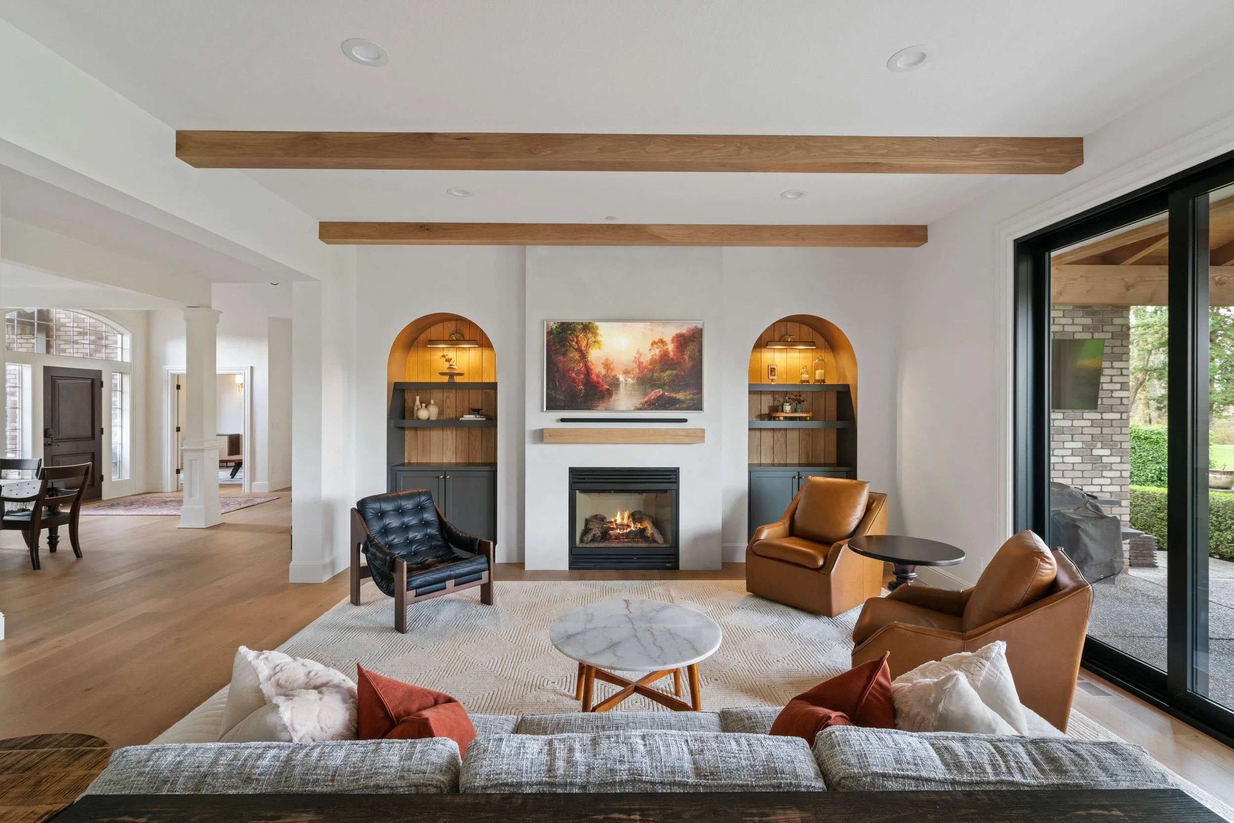 Living room with white walls, wooden ceiling beams, a fireplace, a large window to the right, and modern furniture including a gray sofa with pillows, two leather armchairs, a small round table, and a marble-topped coffee table.