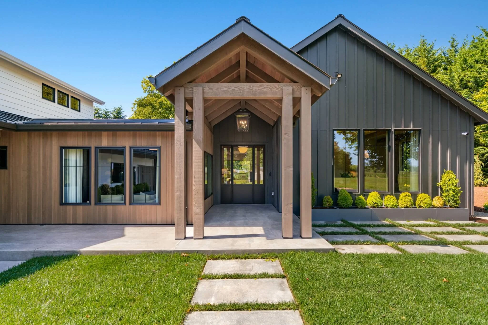 Modern house exterior with wooden and dark gray siding, front porch with wooden supports, concrete pathway, green lawn, and landscaped bushes