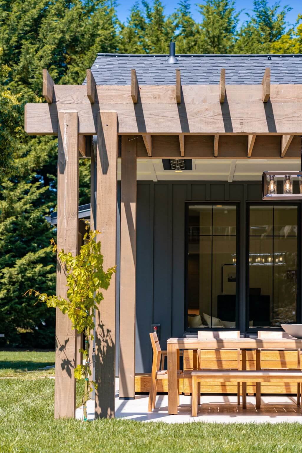 Close-up of a modern house exterior with a wooden pergola, large glass sliding doors, patio furniture, and lush green trees in the background.