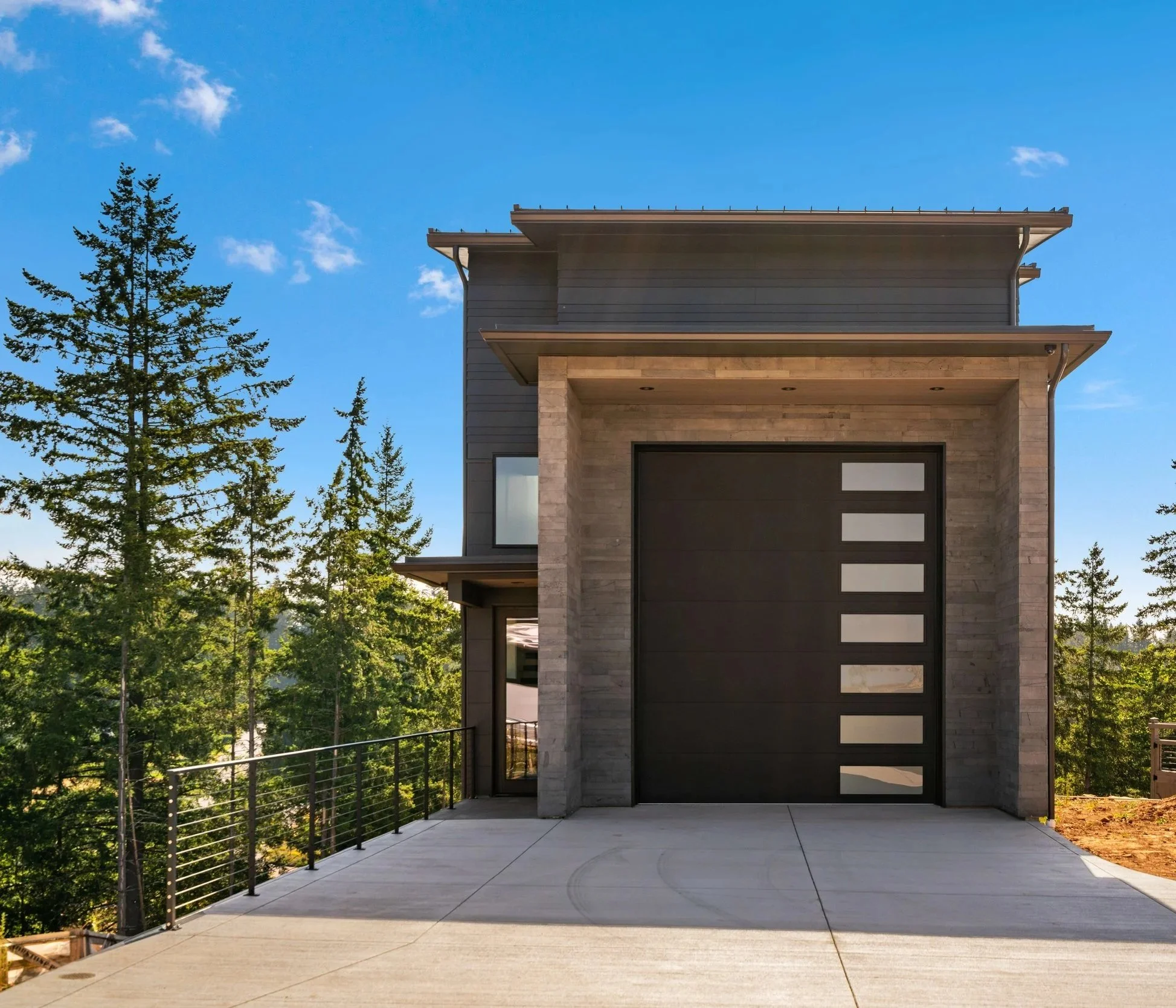 Modern two-story house with a large garage door, concrete driveway, and trees in the background under a blue sky.