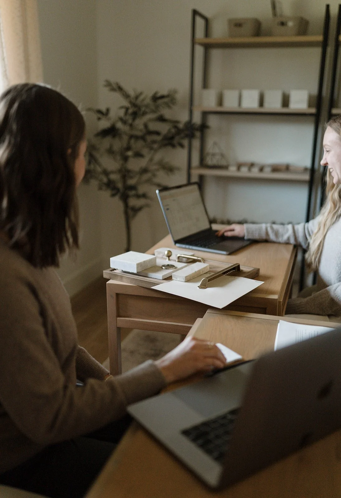 Two designers working at a desk with laptops, notebooks, and office supplies in a cozy, well-lit room.
