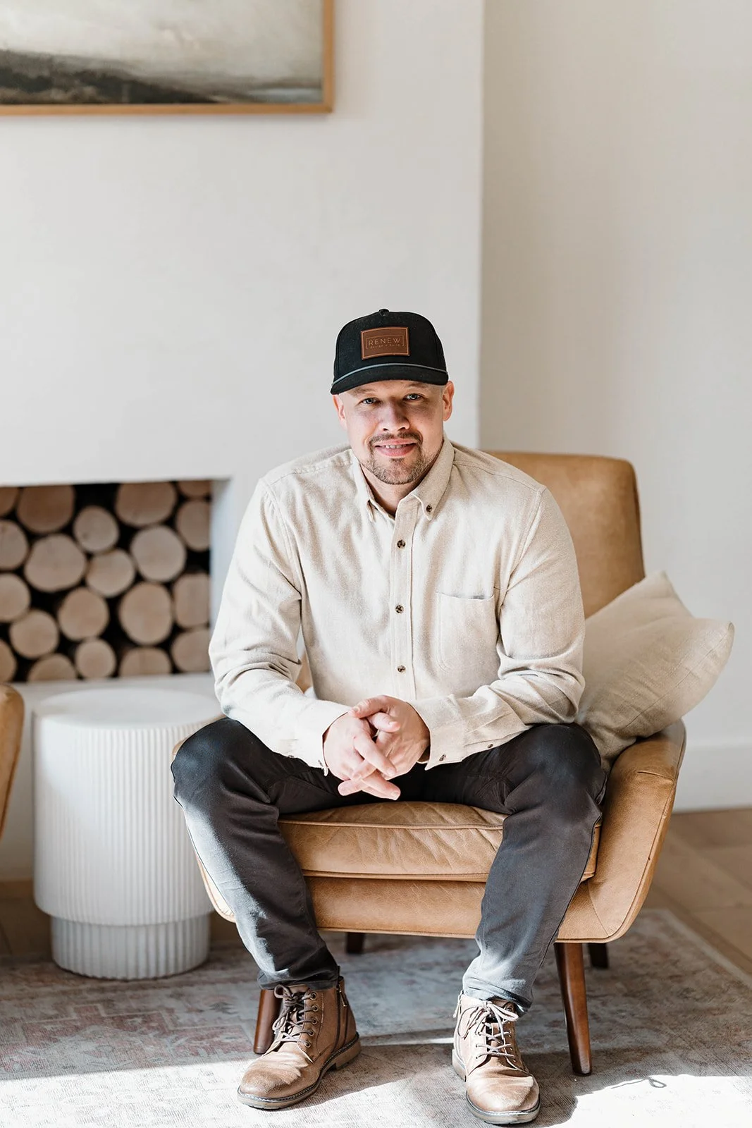 Josh Holmgren, founder of Renew Design + Build, wearing a beige button-up shirt, black jeans, tan work boots, and a black cap sitting on a tan armchair in a minimalist room with a stack of firewood and a side table in the background.