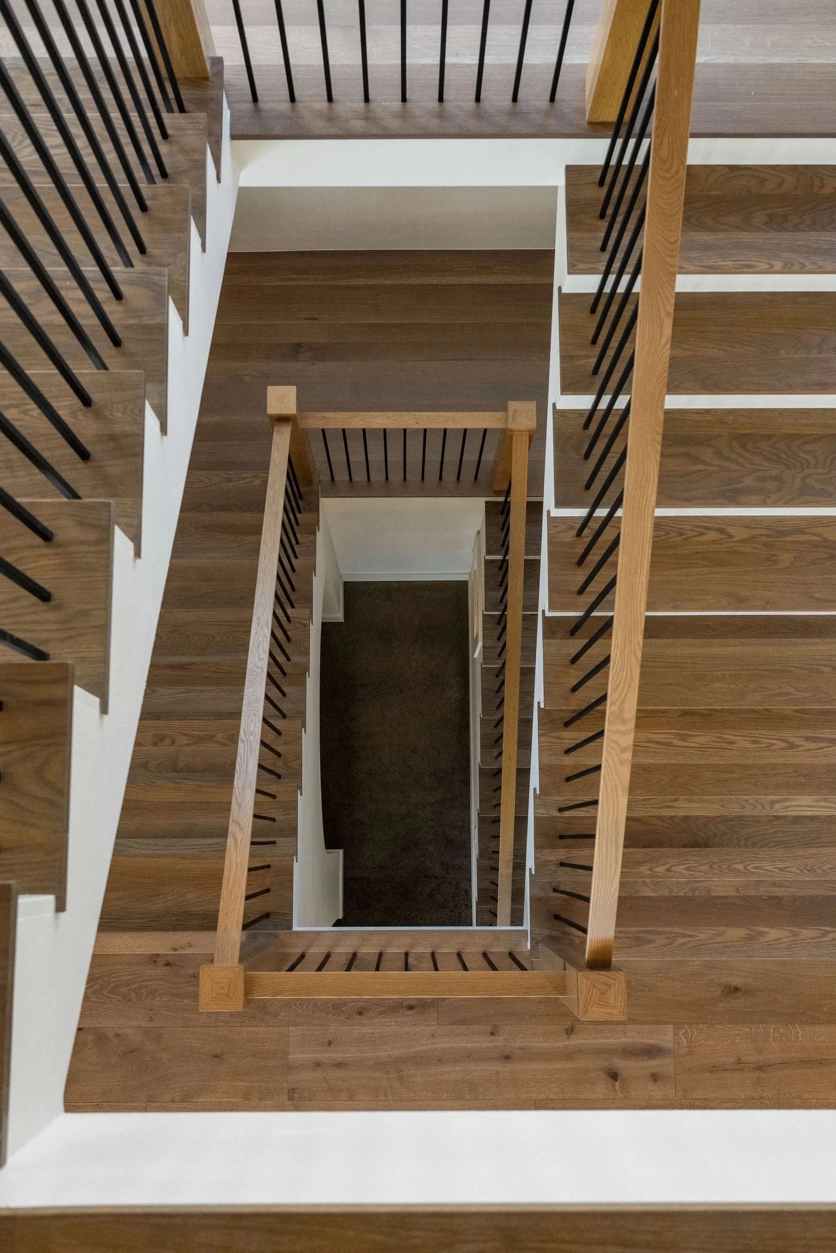 Top view of a staircase with wooden handrails and black balusters, looking down through the center of the staircase.
