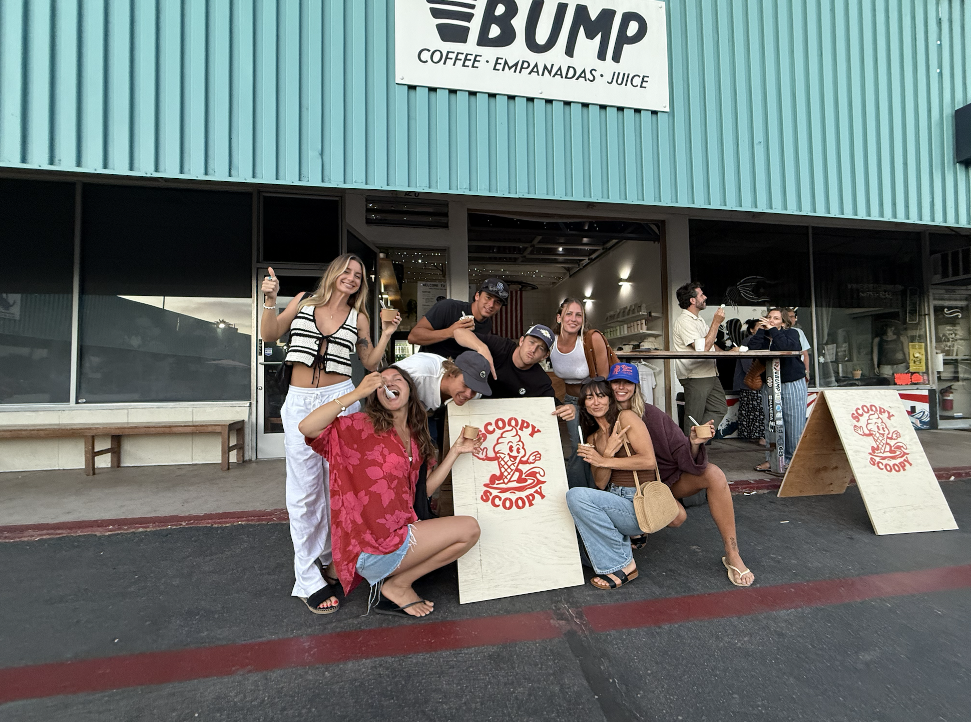 Group of people posing outside Scoopy Scoopy ice cream shop. Some are eating ice cream, smiling, and giving thumbs up. The shop has a signboard with the name and services like coffee, empanadas, and juice.