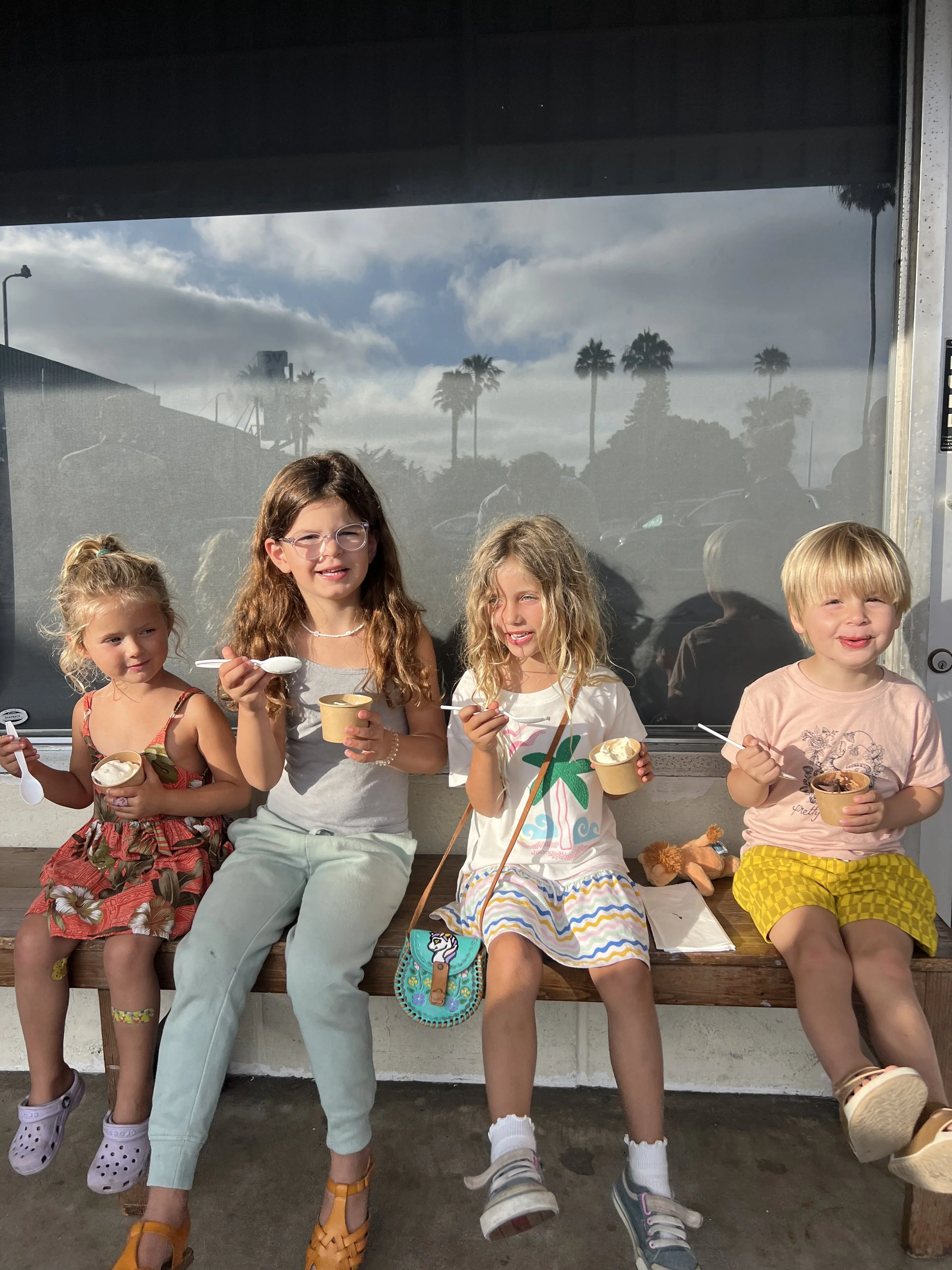 Four children sitting on a wooden bench outside in Cardiff, eating ice cream and smiling, with palm trees and a cloudy sky in the background.
