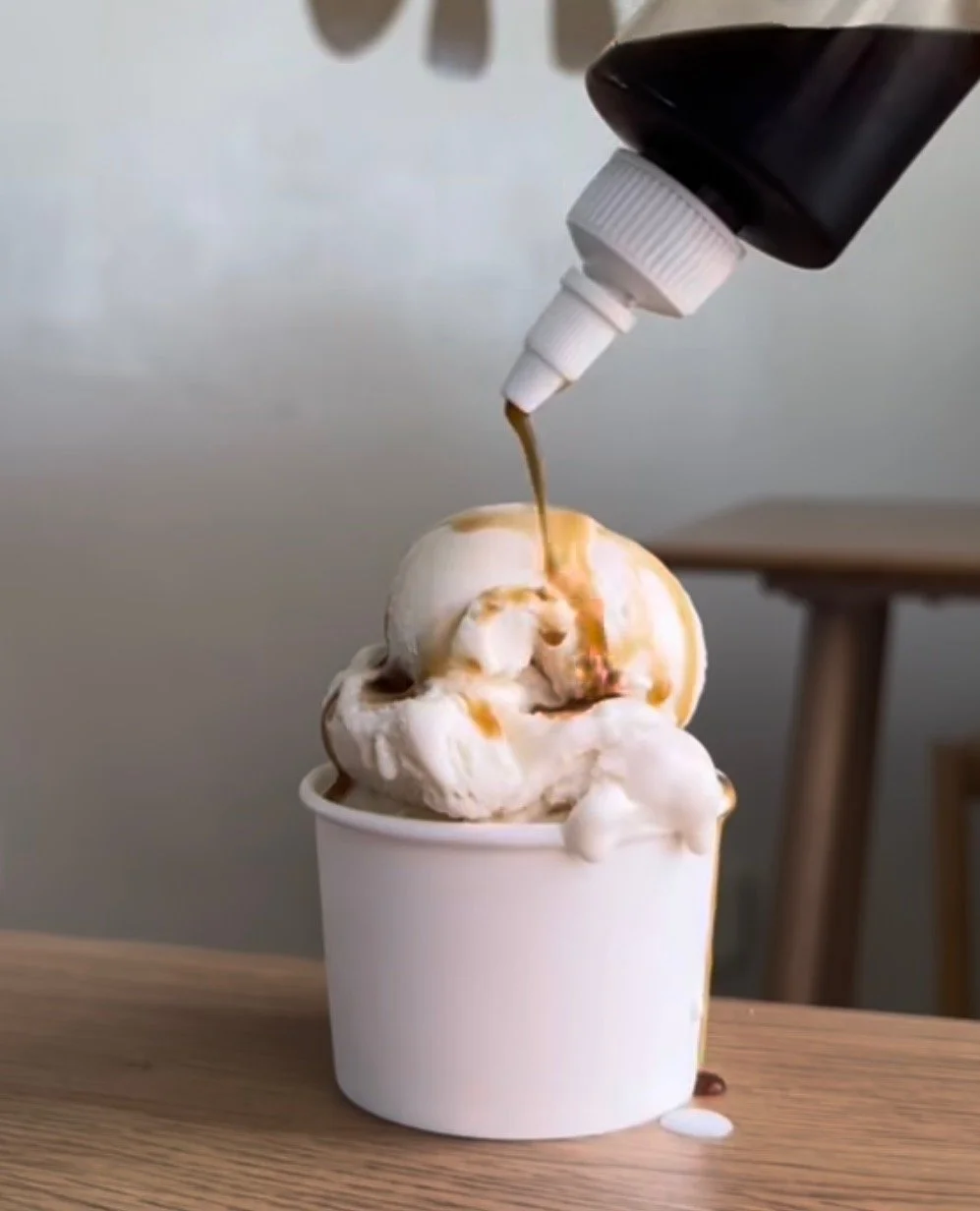 Ice cream in a white cup topped with caramel syrup, with caramel sauce being poured on top.