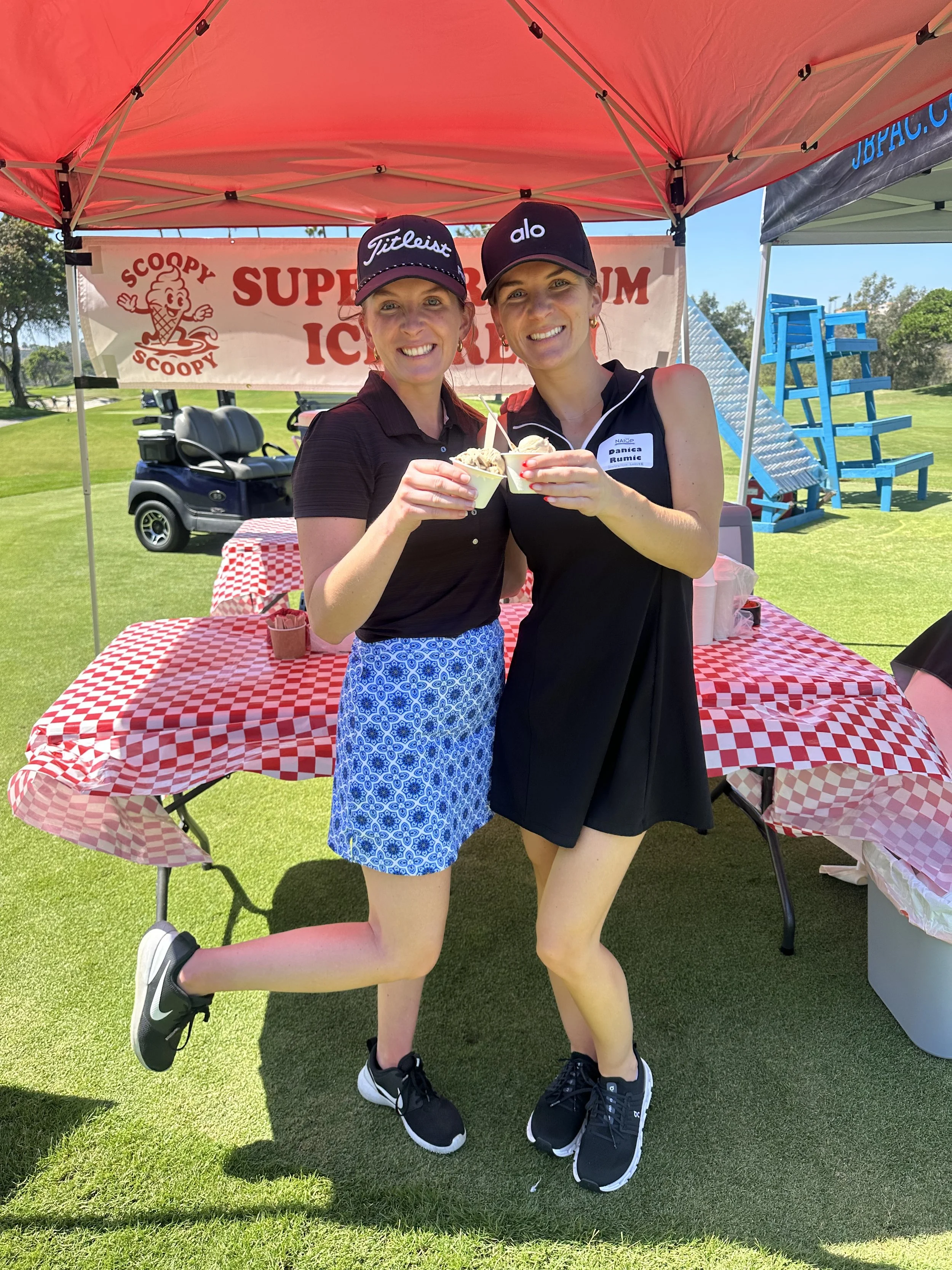 Two women smiling, holding ice cream cups, standing under a red canopy at an outdoor event.