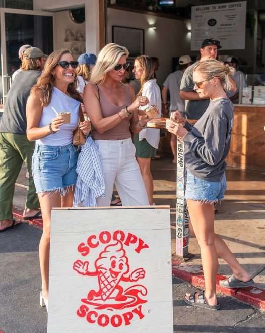 Three women standing outside near a Scoopy Scoopy ice cream stand, holding cups of ice cream and interacting with each other, with other people in line behind them.