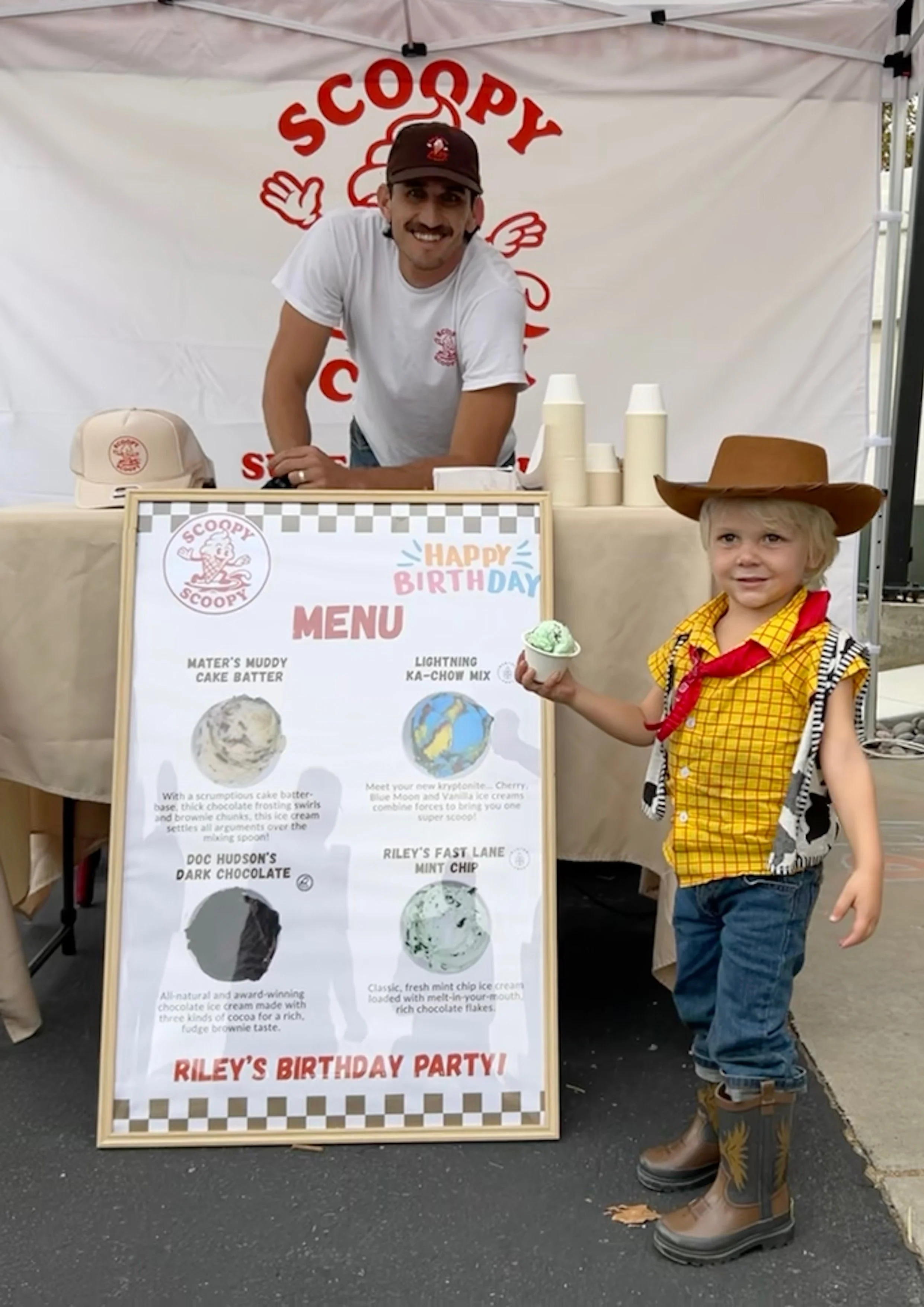 A young boy dressed as a cowboy holding a scoop of green ice cream, standing next to an ice cream menu at a birthday party. A man behind the table smiles, wearing a white t-shirt and a brown cap. The background features a banner with the Scoopy logo 