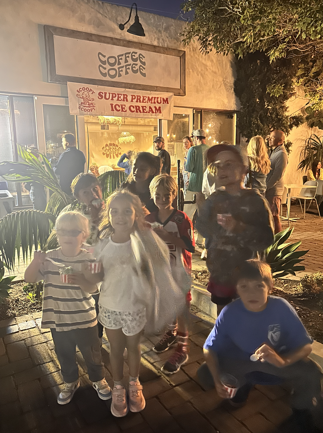 A group of children and adults standing outside an ice cream shop at night in Encinitas, with a sign advertising super premium ice cream. The children are holding bowls or cups of ice cream, and some are smiling at the camera.