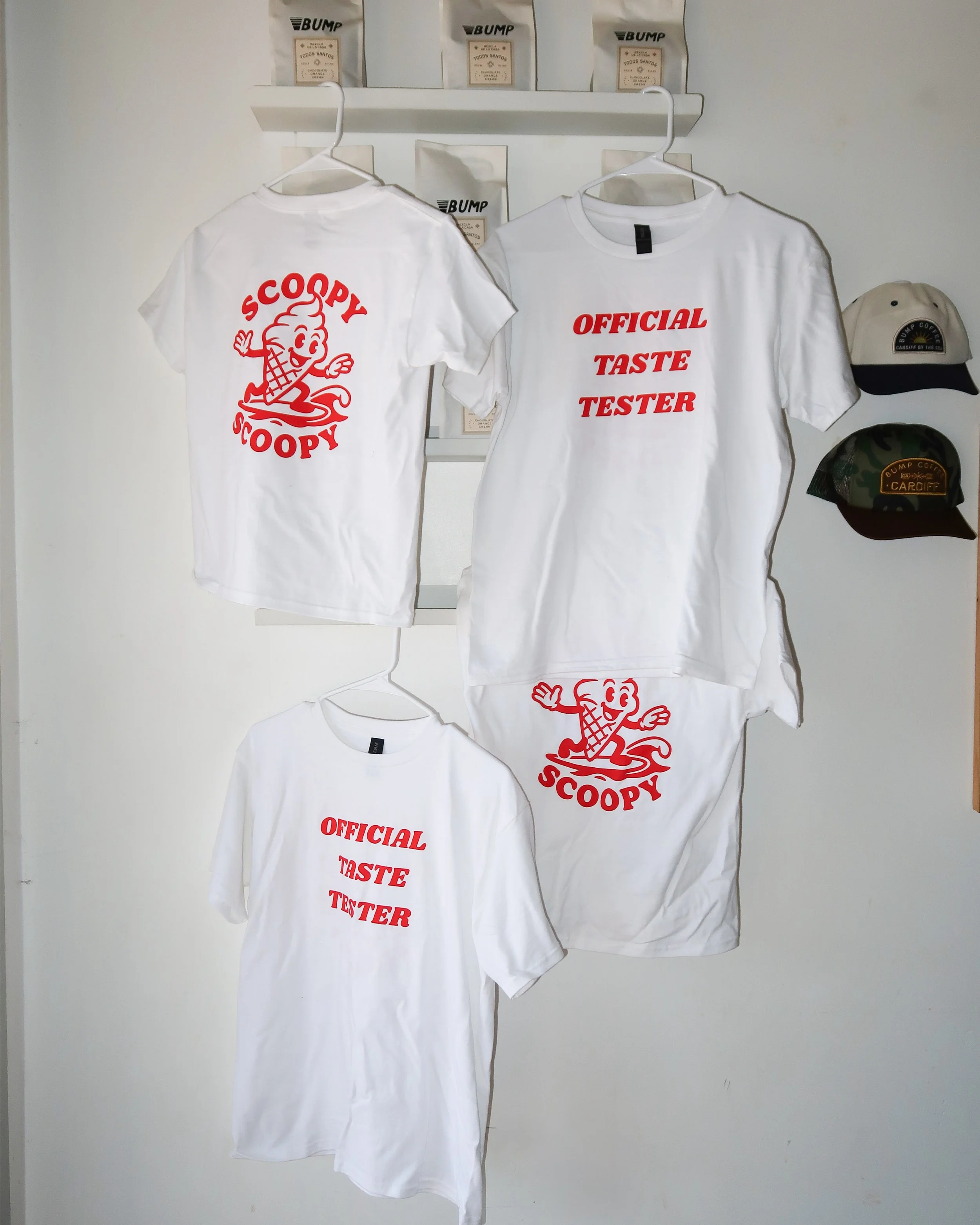 Three white T-shirts with red text and graphics hanging on the wall. Two feature a cartoon ice cream cone character with the words "Scoopy". One shirt says "Official Taste Tester" and the other has the cartoon ice cream cone.