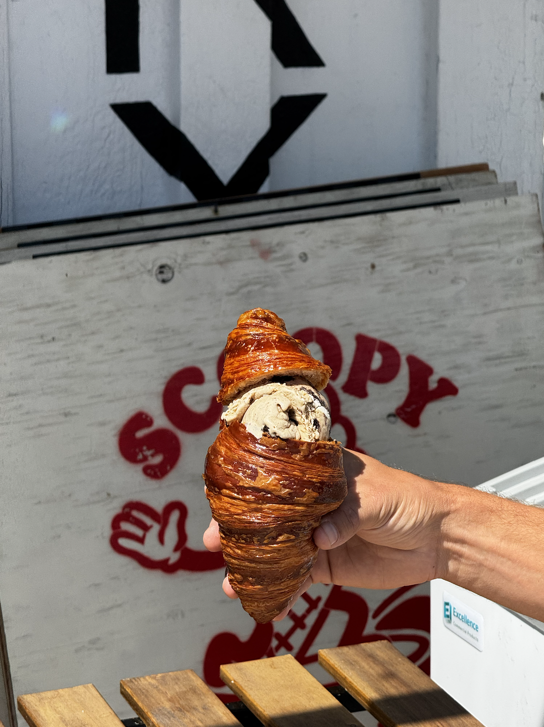 Person holding a croissant filled with ice cream in front of a white wall with red and black graffiti.
