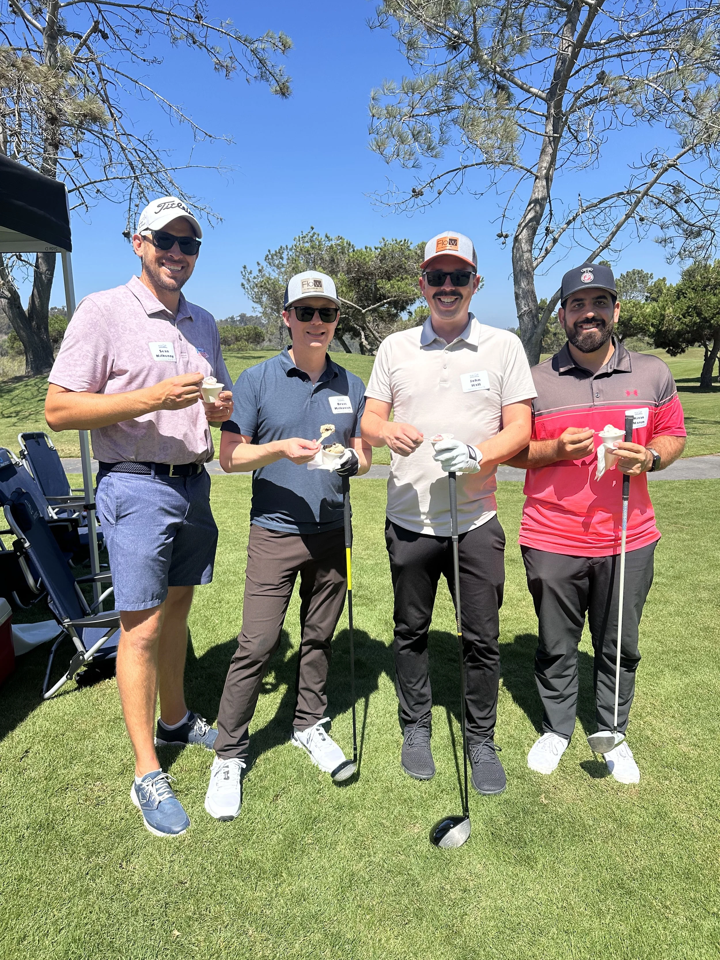 Four men on a golf course, smiling, holding ice cream cups, with golf clubs, under trees on a sunny day.