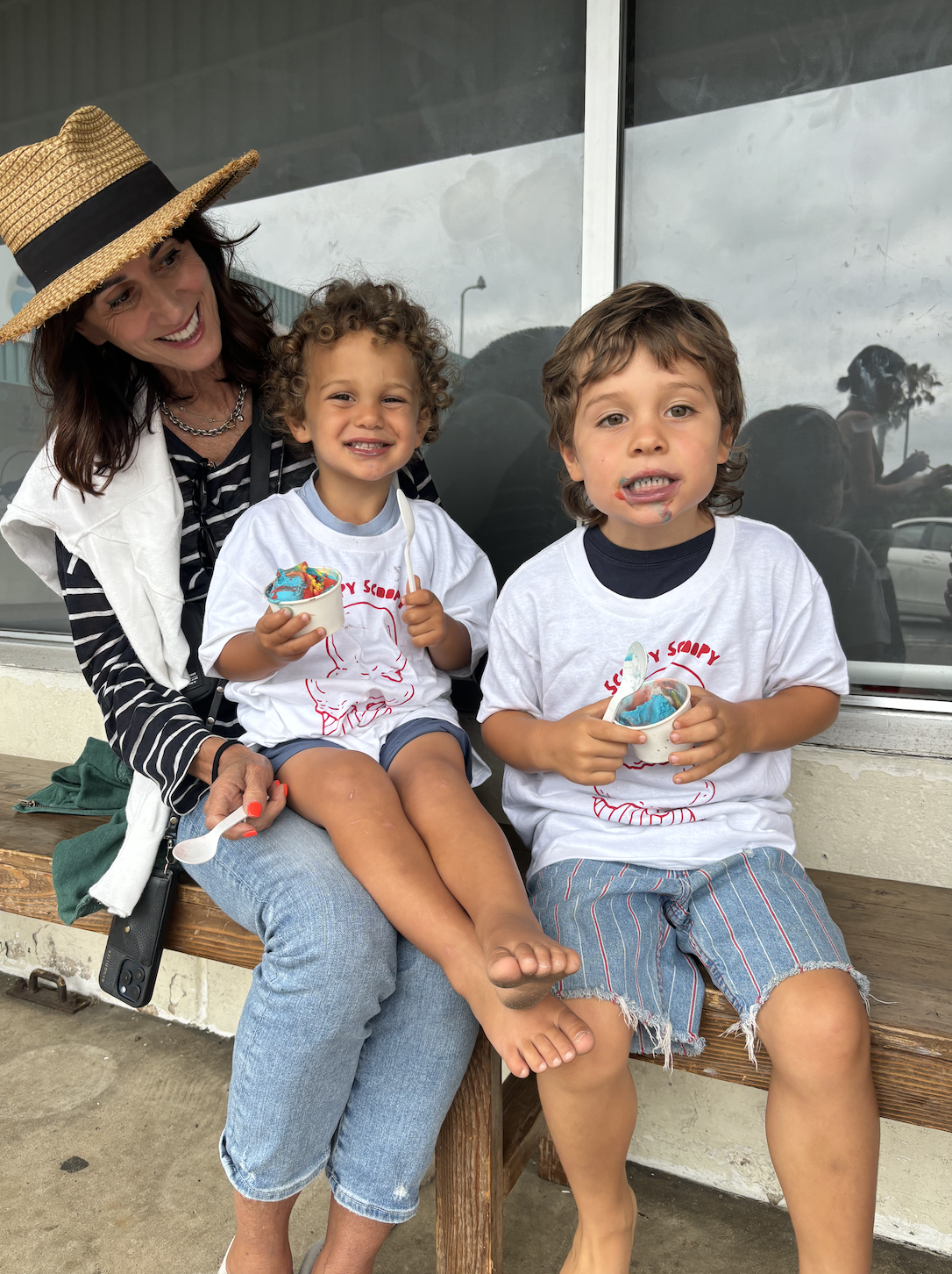 A woman with a straw hat and striped shirt sitting on a wooden bench with two young boys, all holding cups of ice cream, outside on a cloudy day.