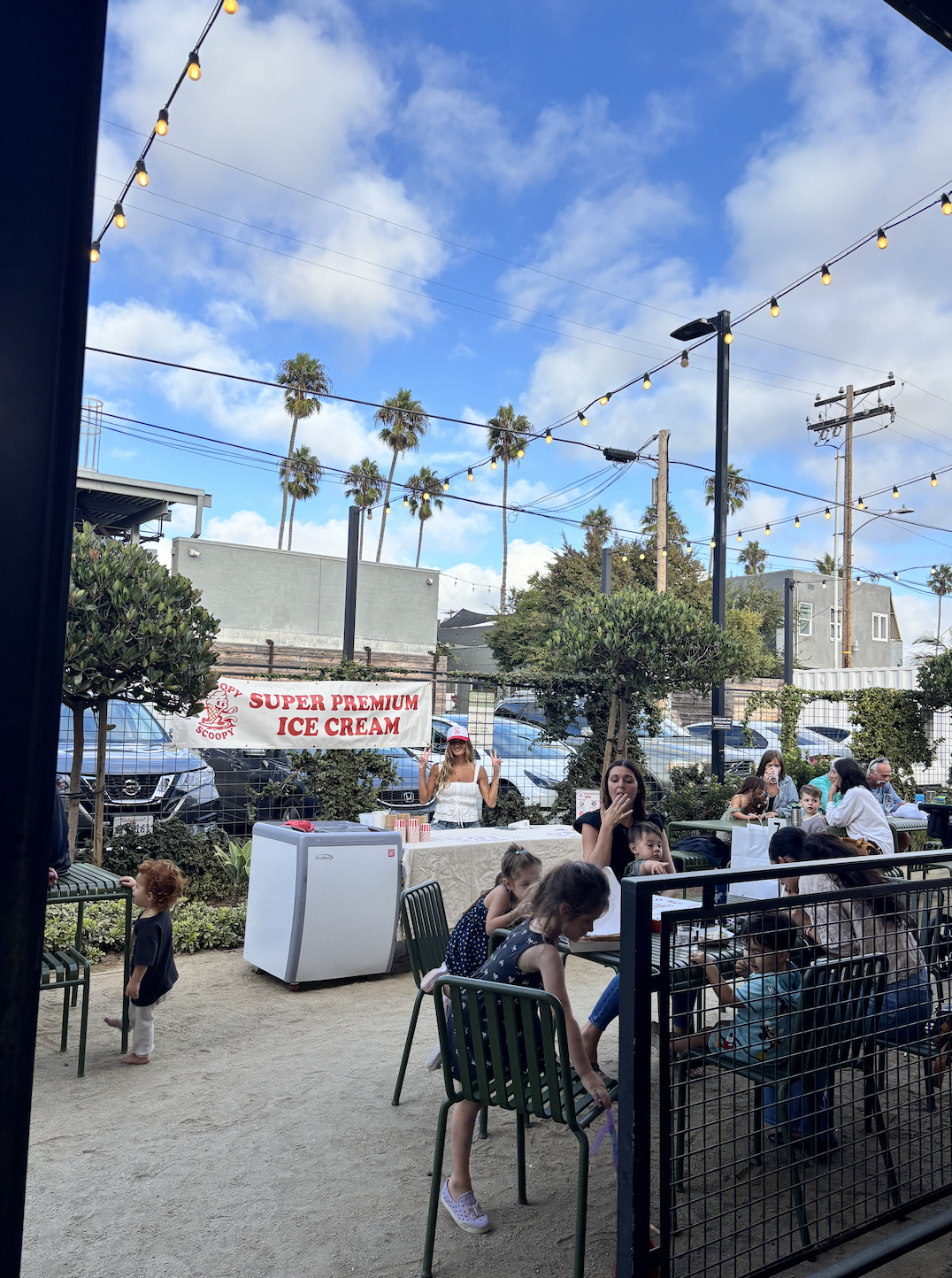People sitting outdoors at a gathering with a banner advertising super premium ice cream, children playing, and a vendor woman in a white top and red cap near the ice cream stand. There are string lights overhead, cars parked in the background, and p