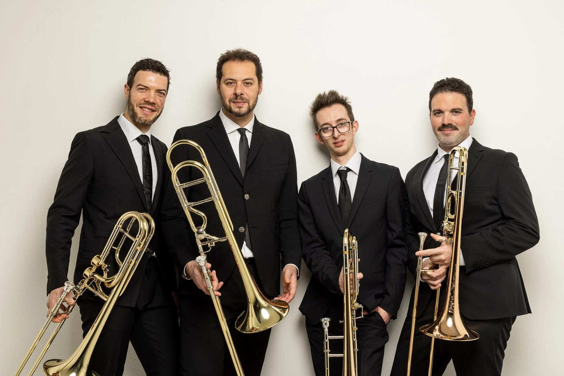 Gruppo di cinque uomini in abiti formali con strumenti musicali a fiato, posizionati contro una parete bianca.